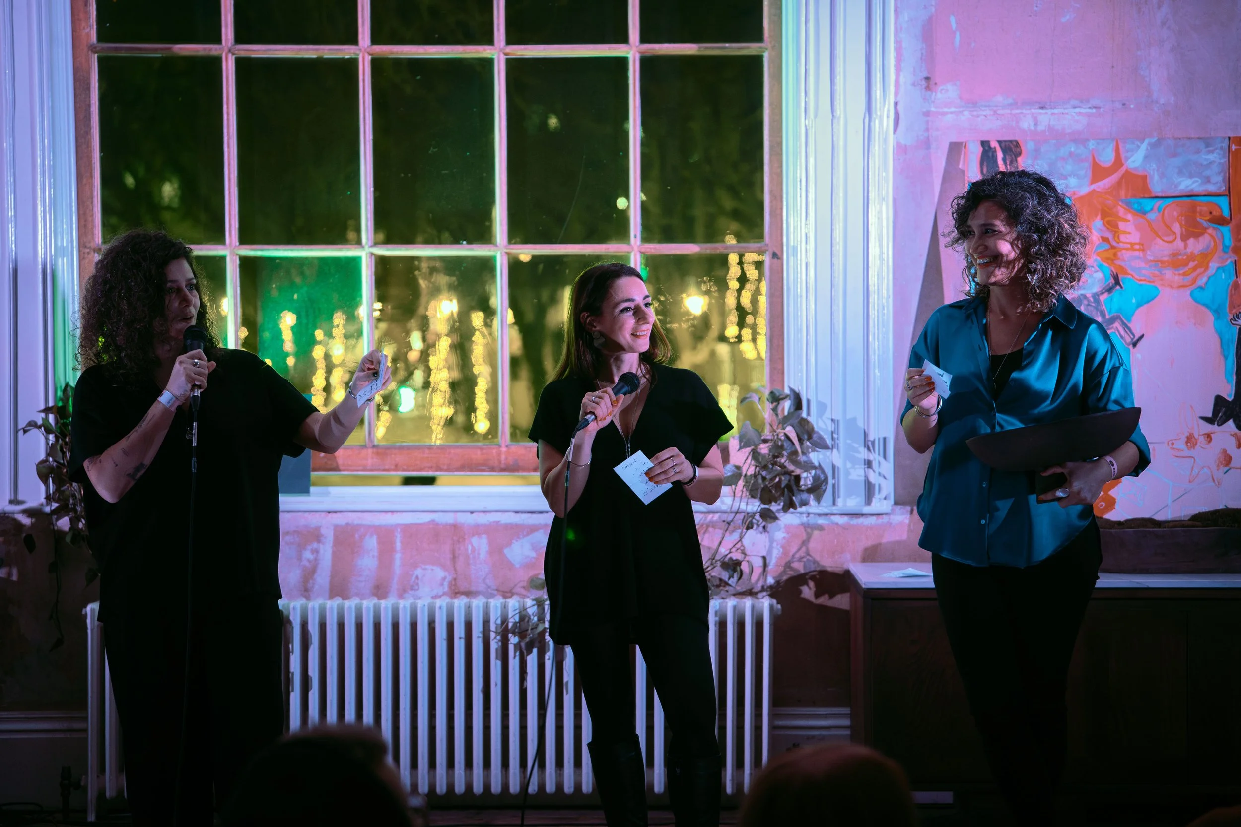 What Does Not founder, Christine, and the rest of the team, Tamara and Mary at RAW uncensored storytelling. The three women speak into microphones at the RAW event with colorful lighting, large windows, and abstract artwork on the wall.