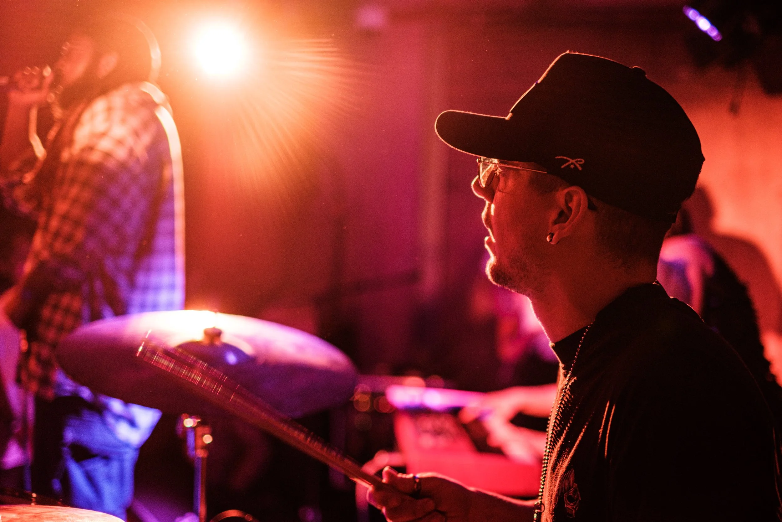 A drummer wearing glasses and a baseball cap playing drums in a colorful, lit performance space at a What Does Not live music night in London.