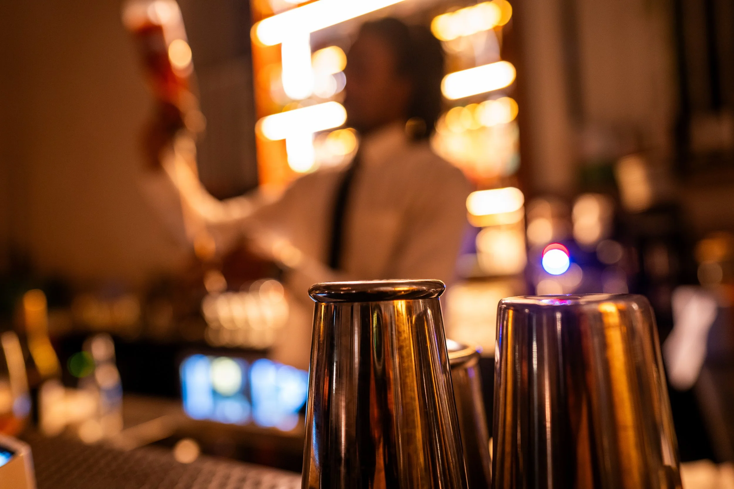 Close-up at RAW uncensored storytelling of shiny metal cocktail shakers on a bar counter with blurred warm-toned lights and glasses in the background.