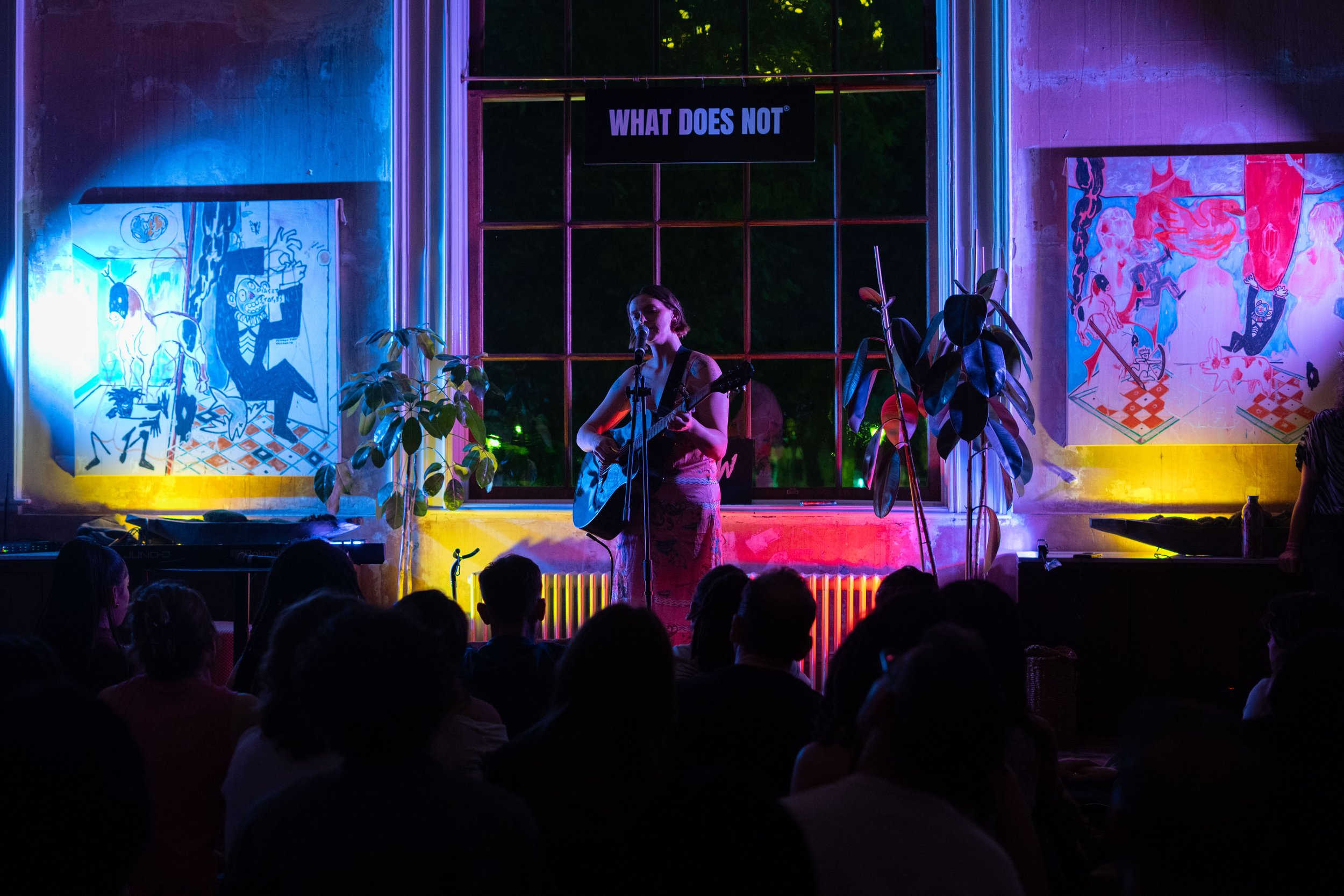 A woman is performing a guitar and microphone set on stage at RAW uncensored storytelling in London, in a dimly lit room with colorful neon lighting, with abstract artwork on the walls and an audience watching.