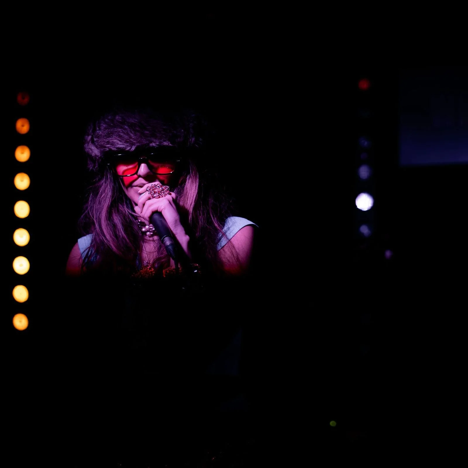 Woman with long hair, sunglasses, jewelry, holding a microphone, on a dark stage with colorful lights at a What Does Not live music night