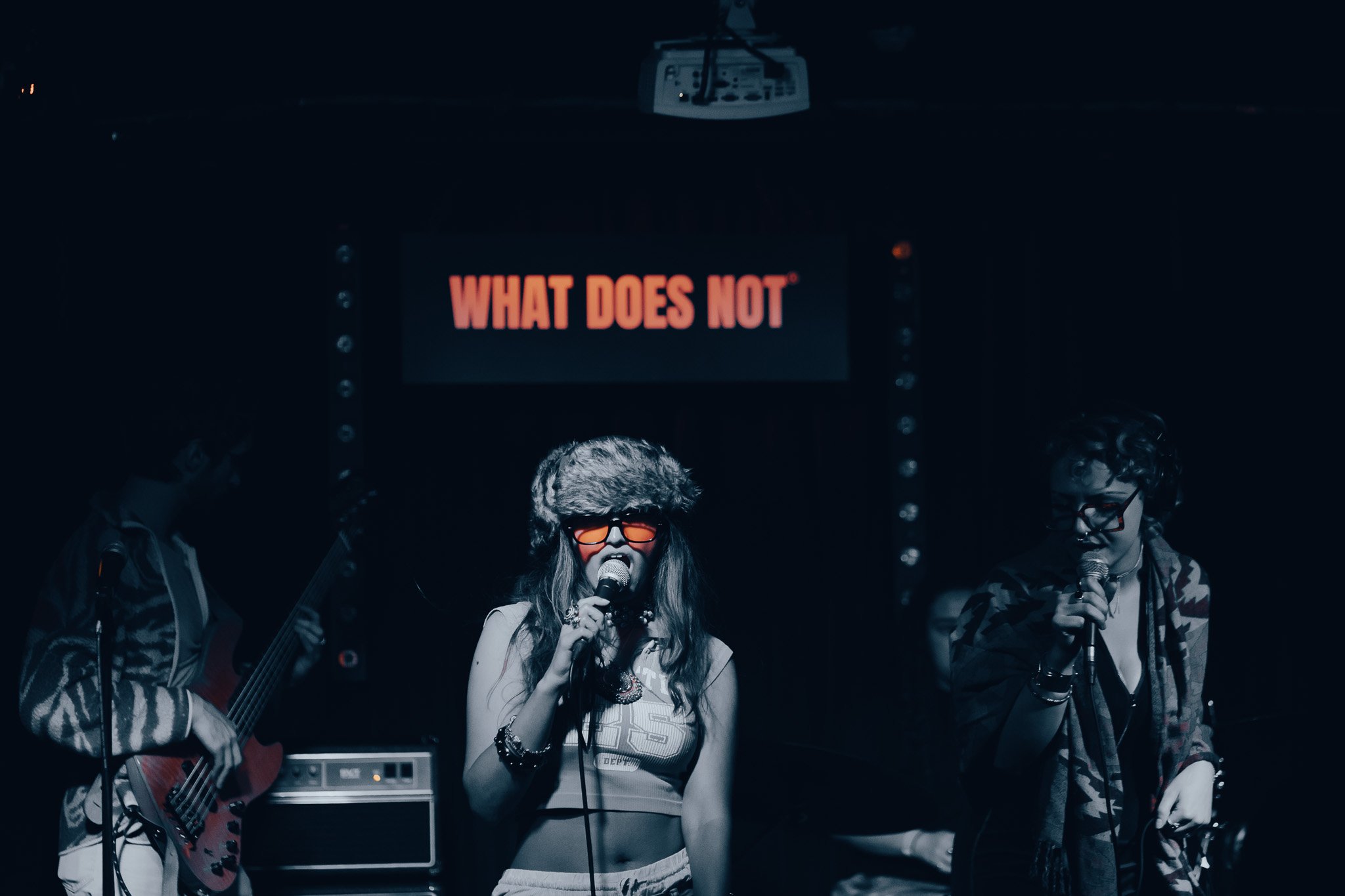 A group of three musicians performing at a What Does Not open jam / music improv night, one woman singing into a microphone and two men playing guitars, with a screen behind them displaying the words 'What Does Not' in bright red letters.