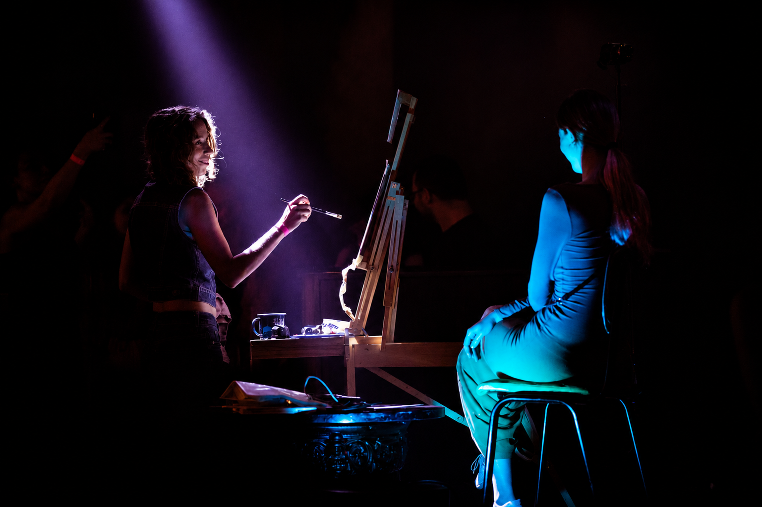 Live painting at a What Does Not event where a woman with brown hair sitting on a stool, is having her portrait painted by a female artist with dark hair holding a paintbrush.
