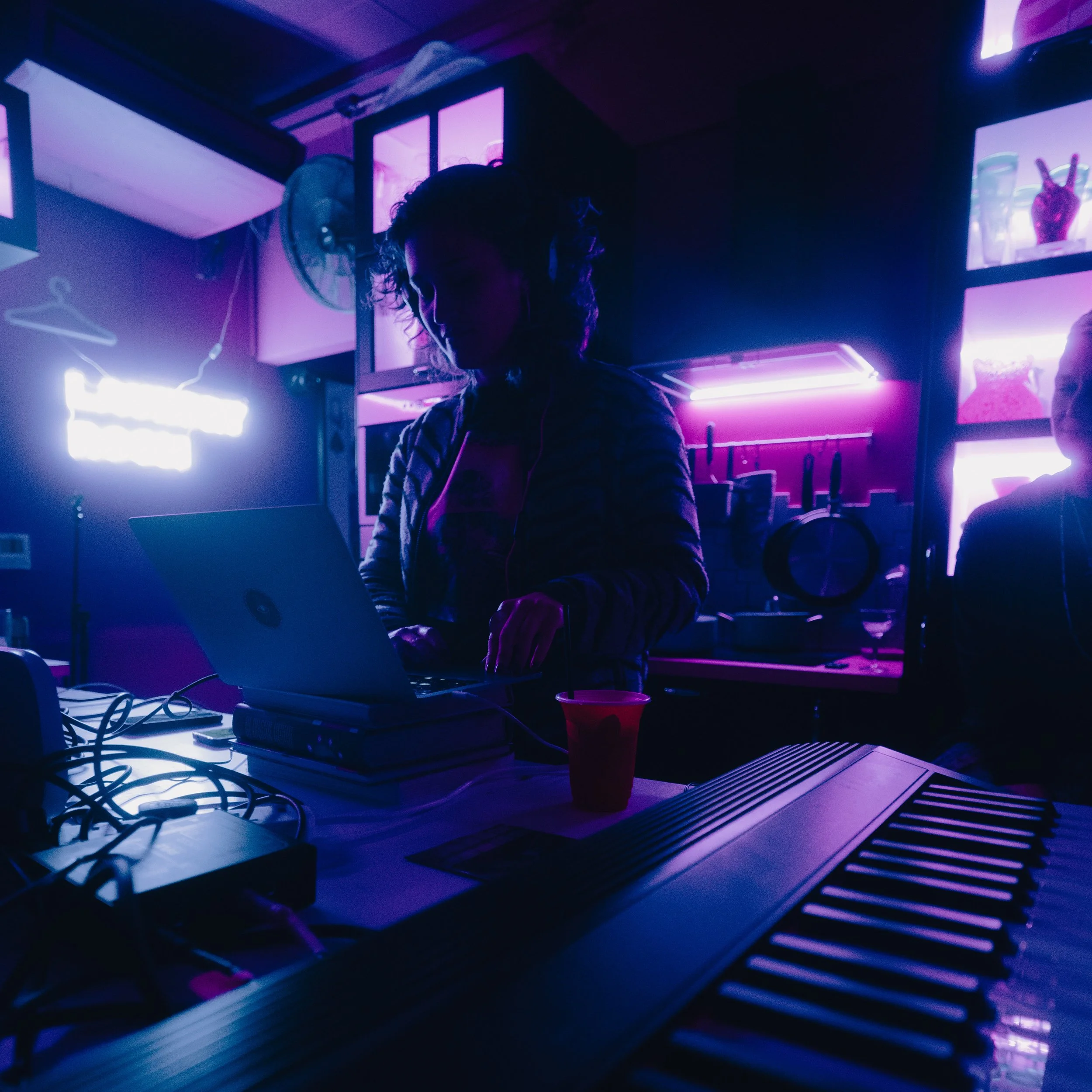 Mary from What Does Not DJs using a laptop at a What Does Not house party in a dimly lit room illuminated by colorful neon lights, with multiple lights on the wall behind her