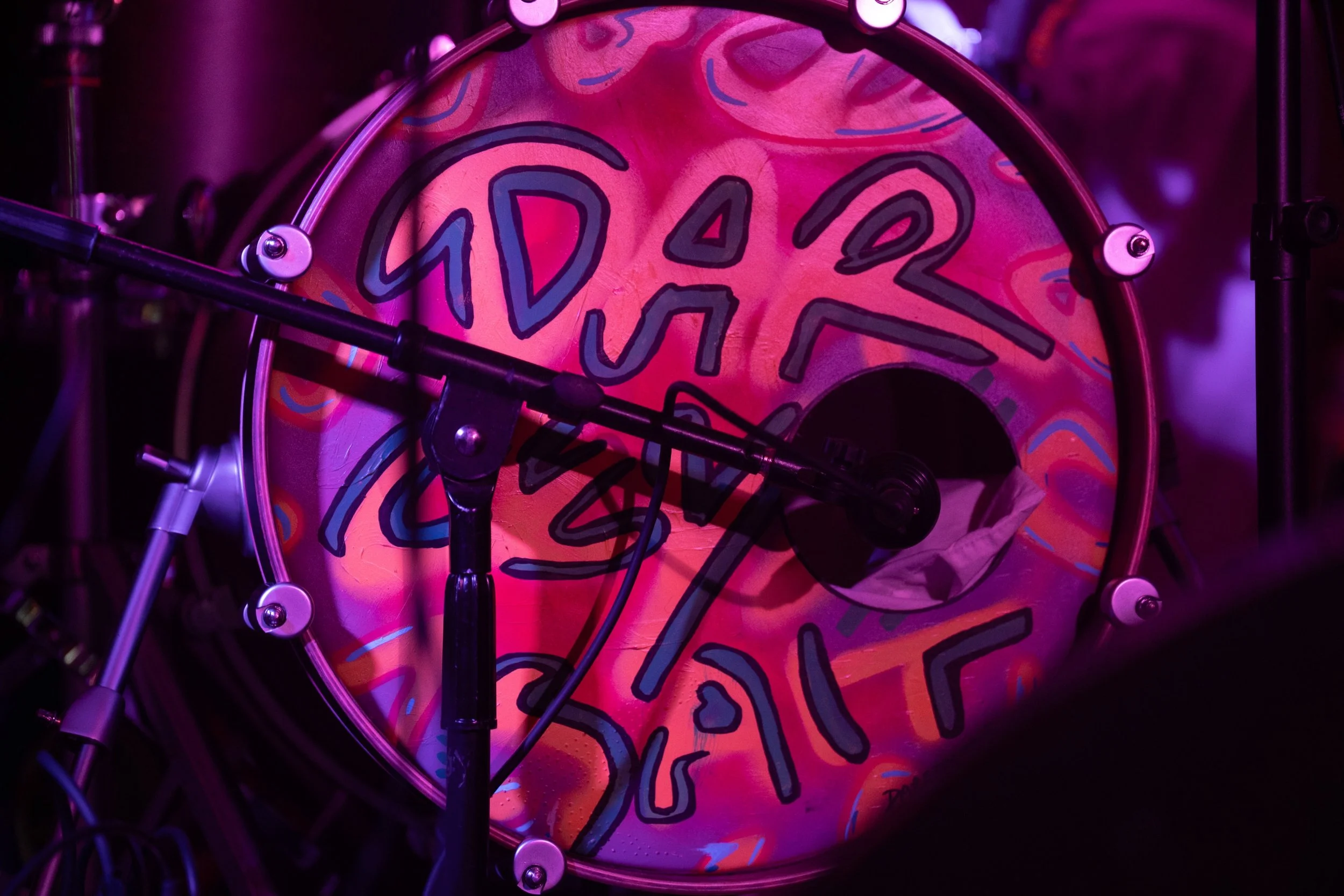 Close-up of a colorful drum with abstract designs in pink, purple, and blue, surrounded by drum hardware and purple lighting.