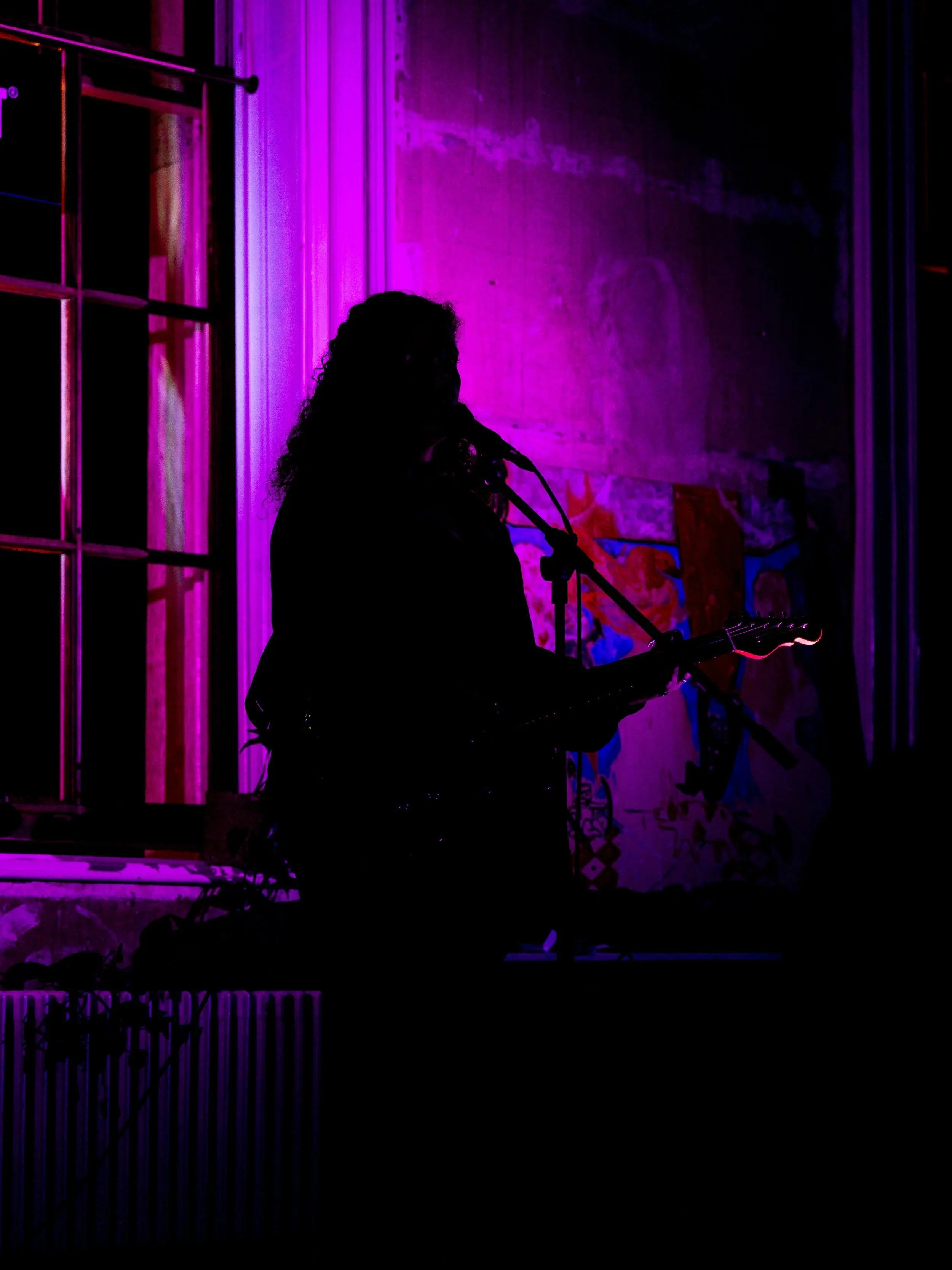 An emerging musician performs at RAW uncensored storytelling, playing an electric guitar in a dimly lit room with purple and pink neon lighting, standing near a wall with graffiti art.