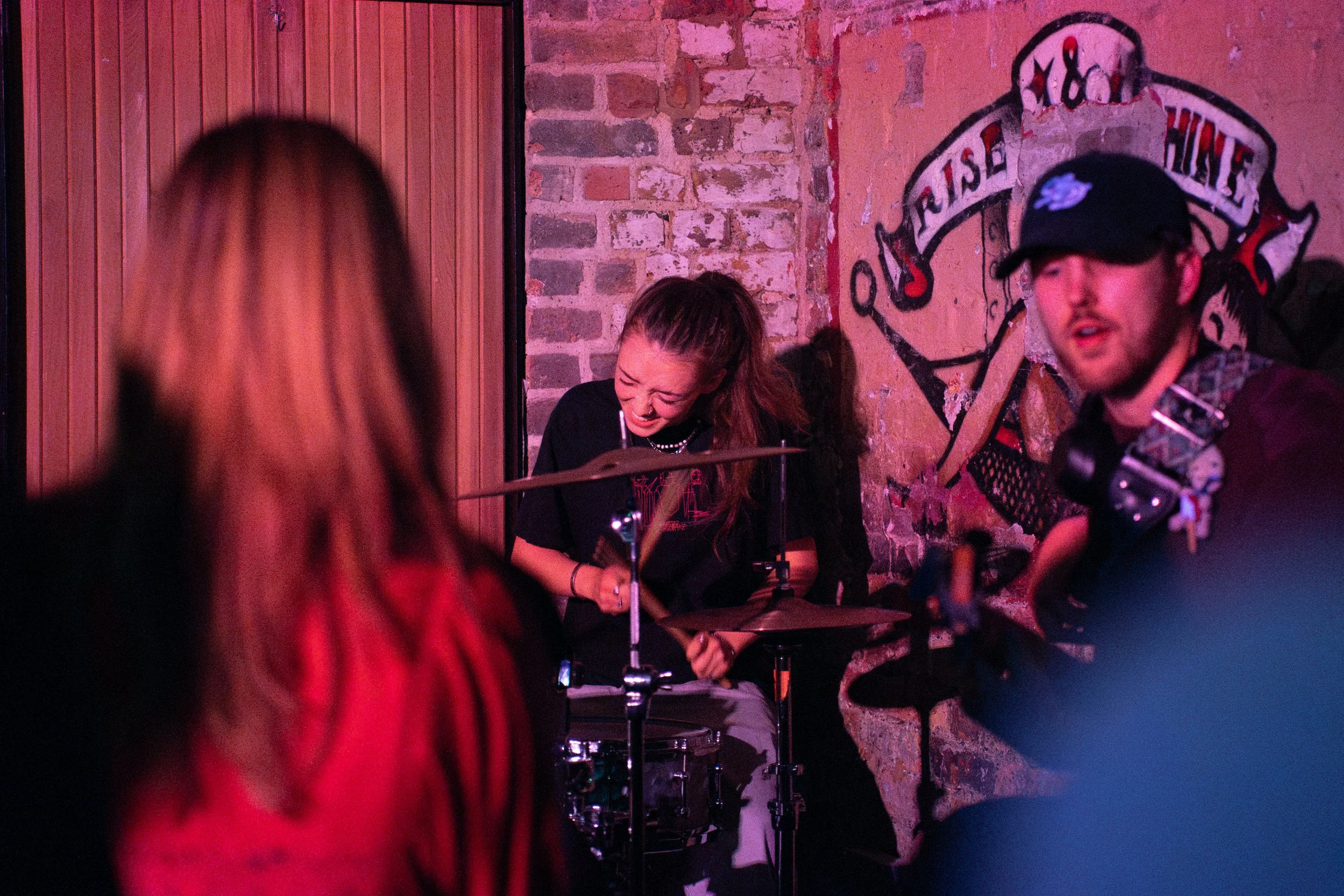 A woman playing drums and singing during a live music performance in a cozy venue with brick and wood walls, featuring a large vintage-style logo on the wall behind.