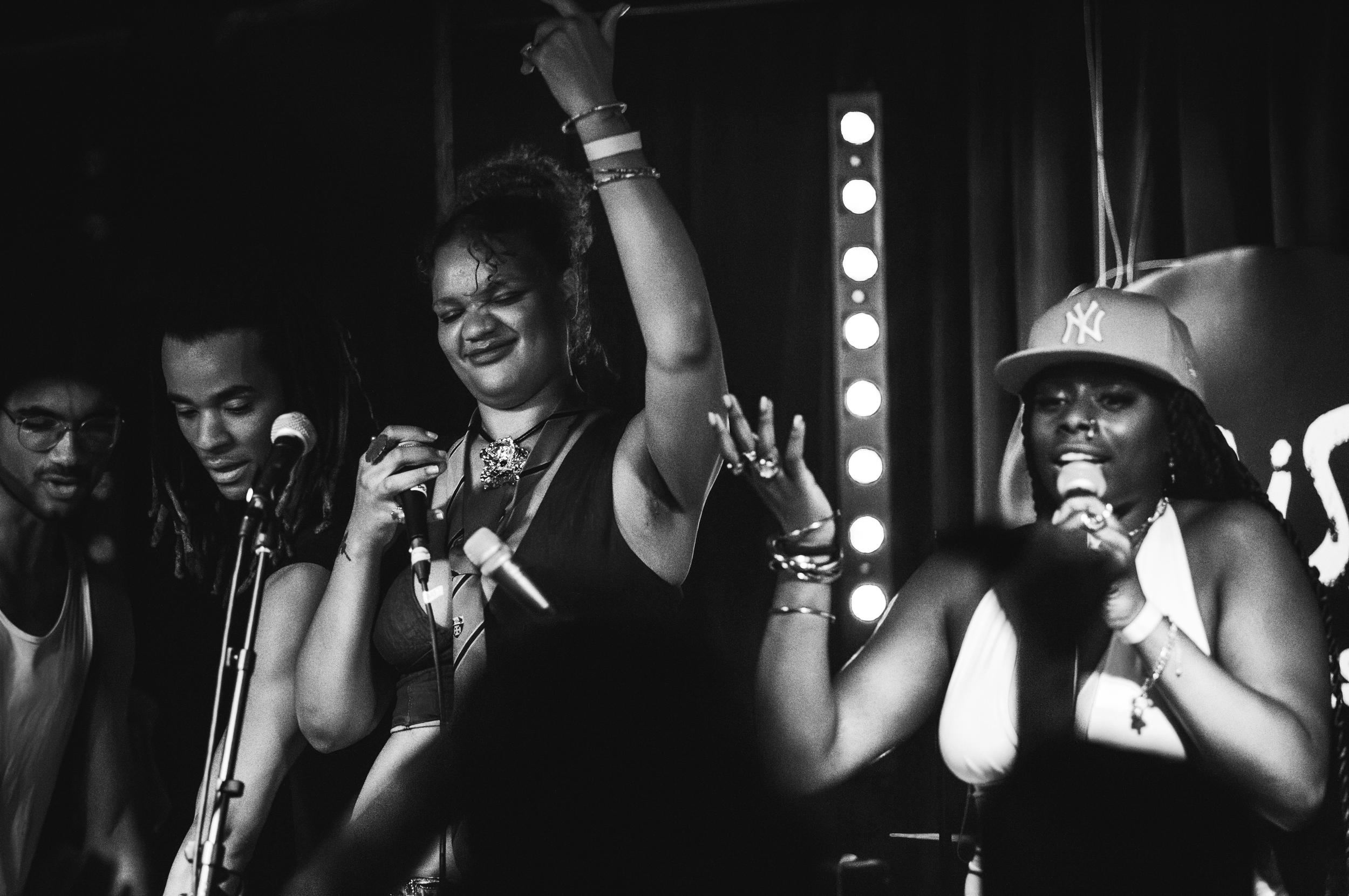 A group of diverse women singing or speaking into microphones on a stage, with one woman raising her arm and all appearing to be joyful, in a black-and-white photo.