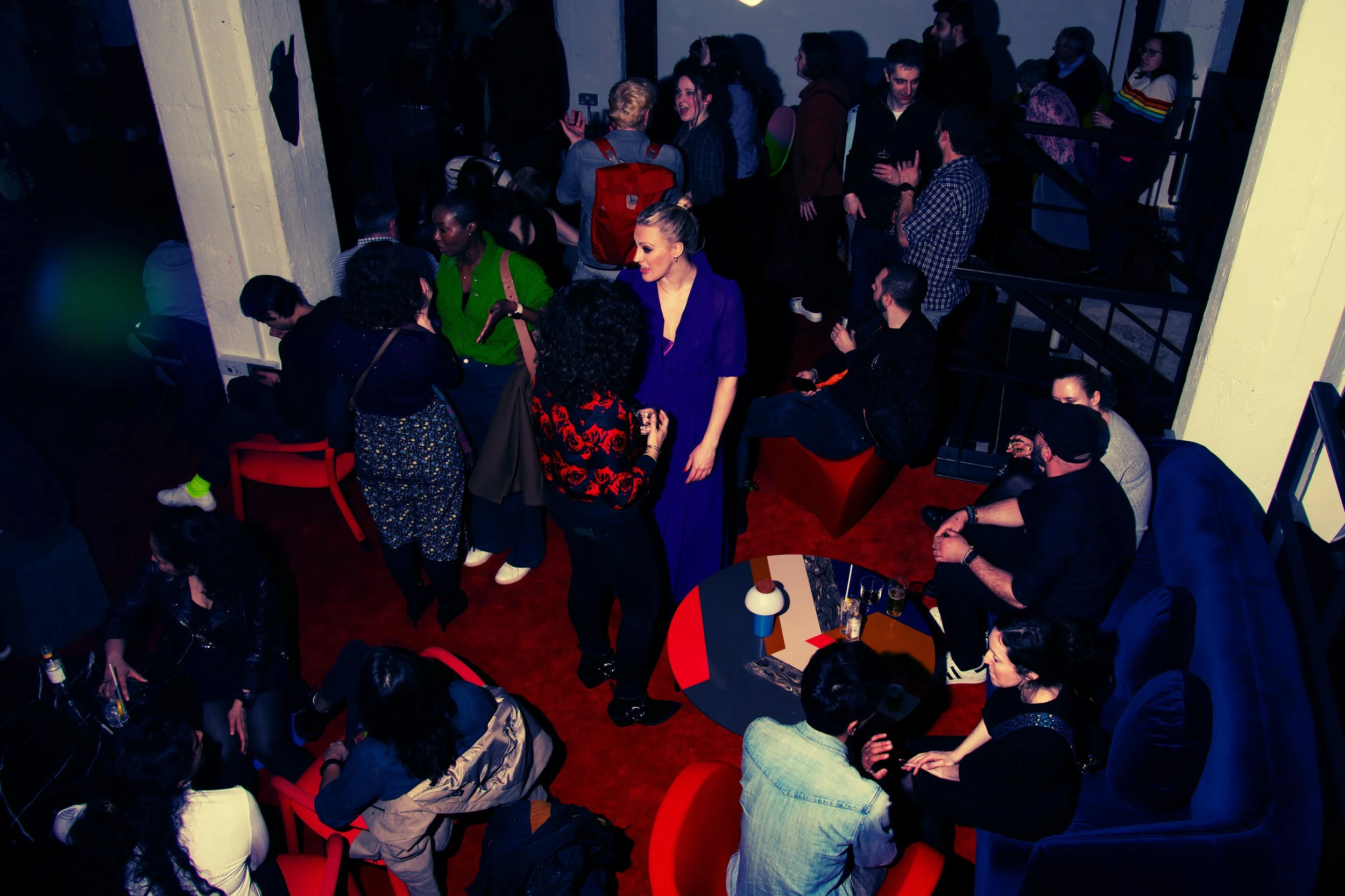 People socializing at a lively gathering called RAW uncensored storytelling, with some standing, chatting, and holding drinks. The room has a dark ambiance with colored lighting and modern furniture.
