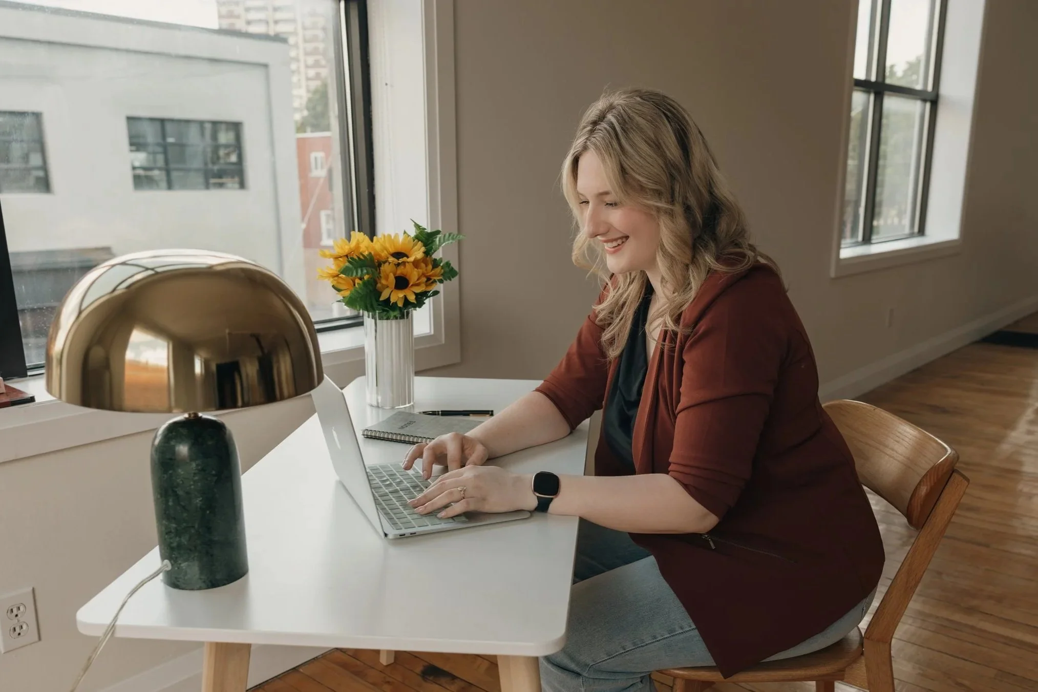 A woman with blonde curly hair sitting at a white table in a modern apartment, working on a laptop, smiling. There is a vase with yellow sunflowers on the windowsill, and a gold-colored lamp on the table. Large windows in the background reveal a cityscape.