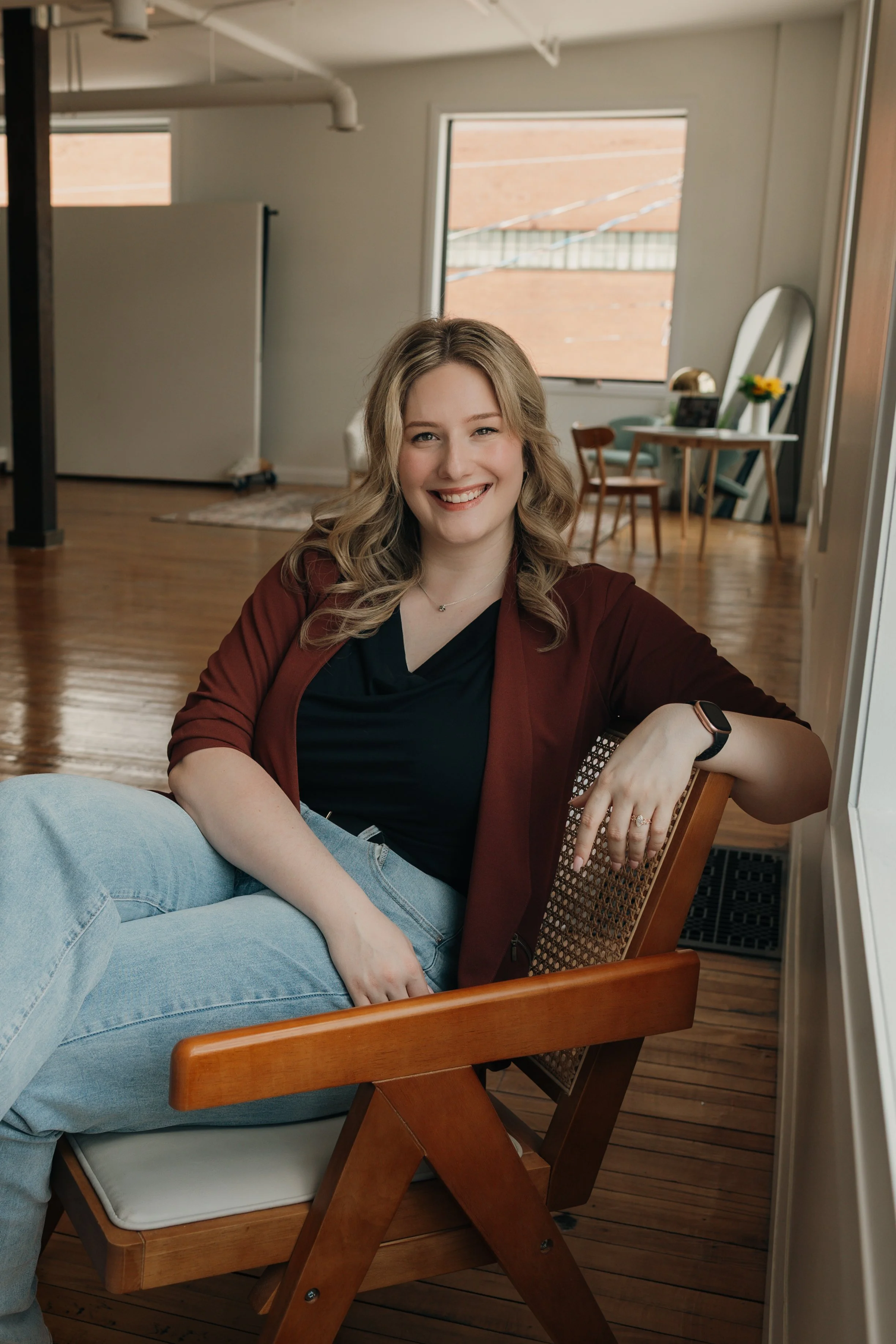 A young woman with curly blonde hair, wearing a burgundy jacket and black top, smiling and sitting on a wooden chair in a cozy room with hardwood floors, a mirror, and a table with flowers in the background.