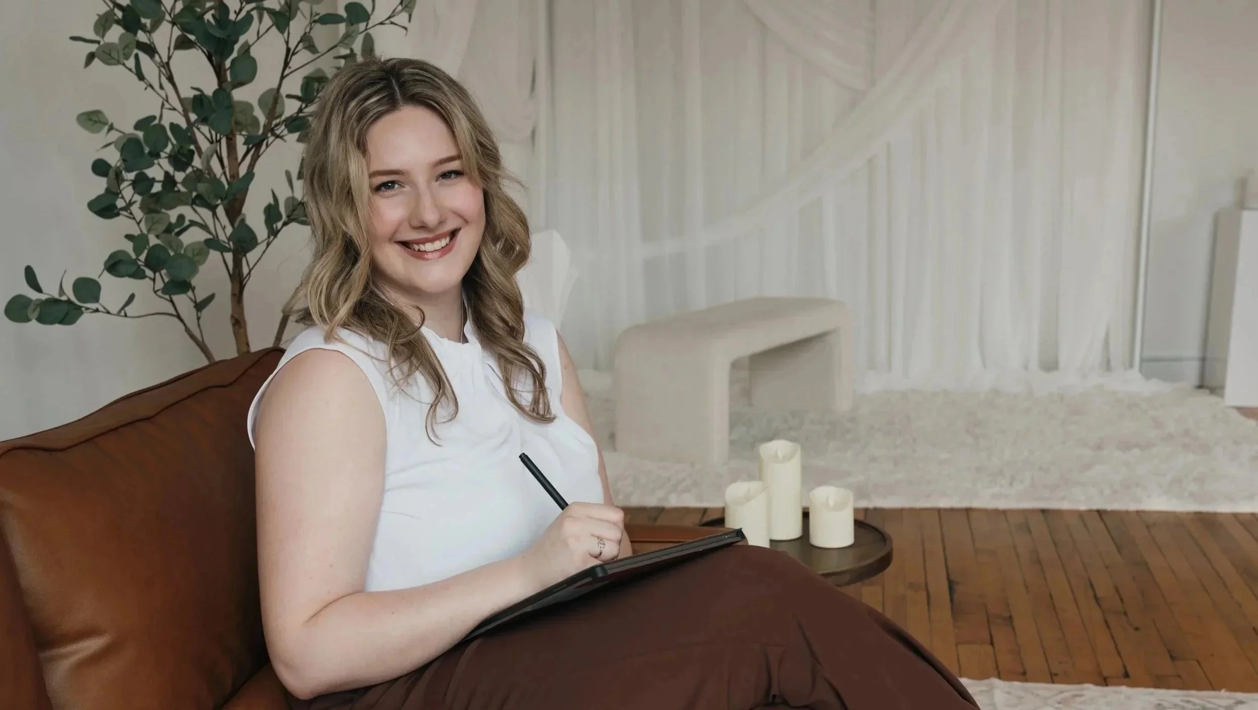 A woman with blonde hair is sitting on a brown couch, smiling, and holding a pen and notebook, with three white candles on a tray beside her. There is a leafy plant behind her, and the background features a white staircase and light-colored walls.