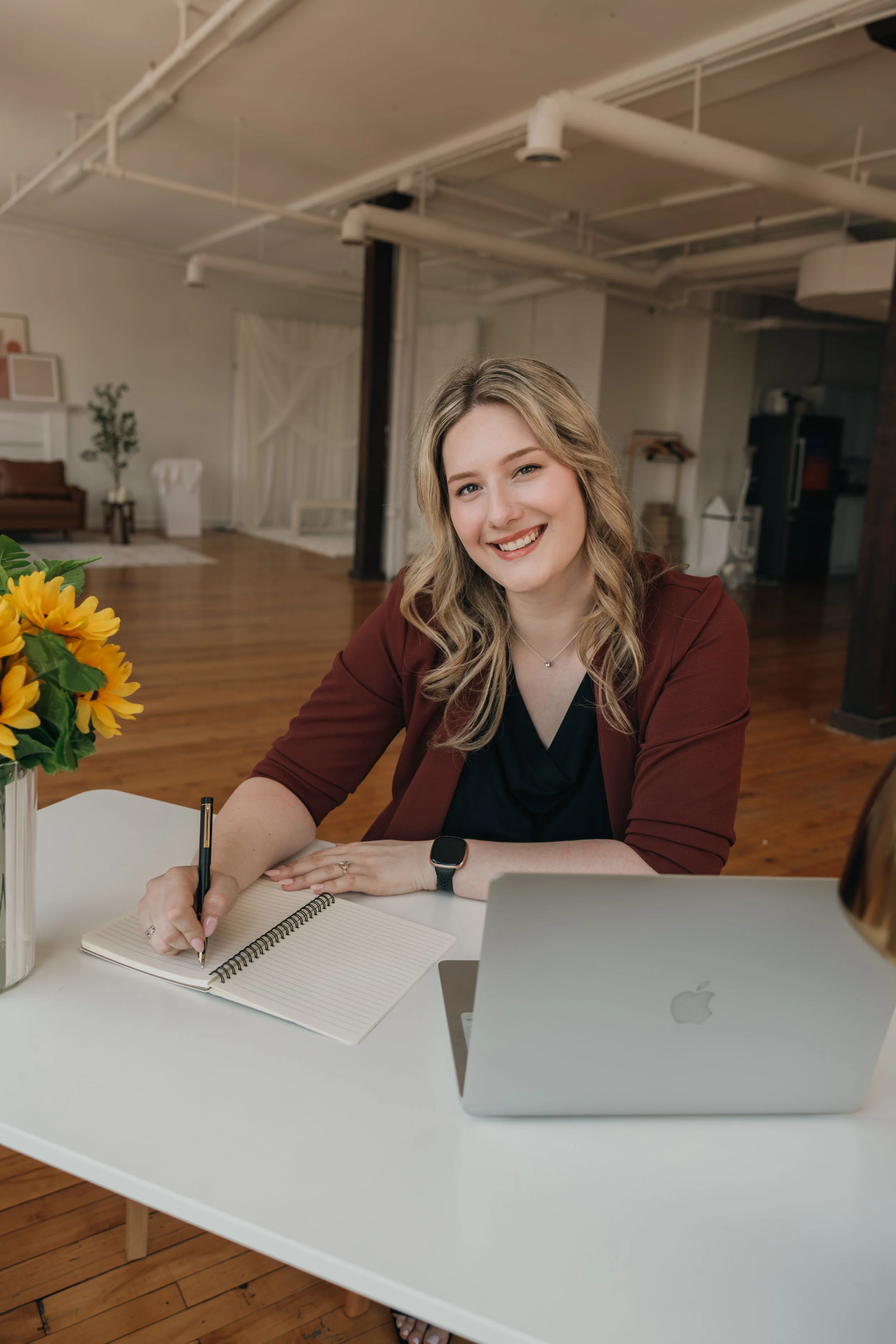 A woman with blonde hair and a friendly smile, wearing a maroon blazer and black top, sitting at a white table in a modern, open-concept office space. She is writing in a spiral notebook, with a laptop displaying the Apple logo, a vase of sunflowers, and a pen on the table. The background features wooden flooring, white walls, and artwork.