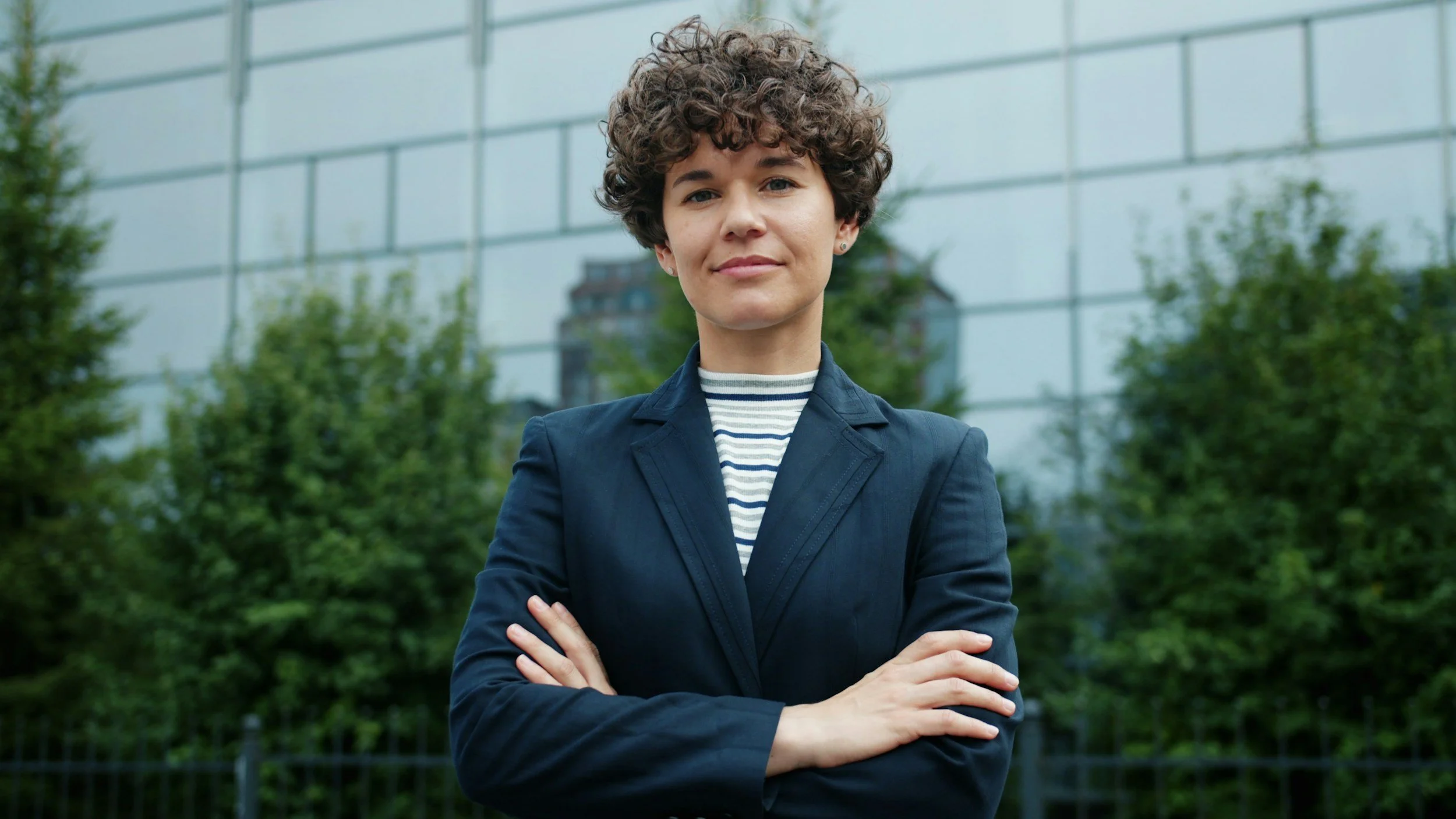 A woman with short, curly hair and light skin standing outdoors with her arms crossed, wearing a navy blazer and striped shirt, in front of a modern glass building and green trees.