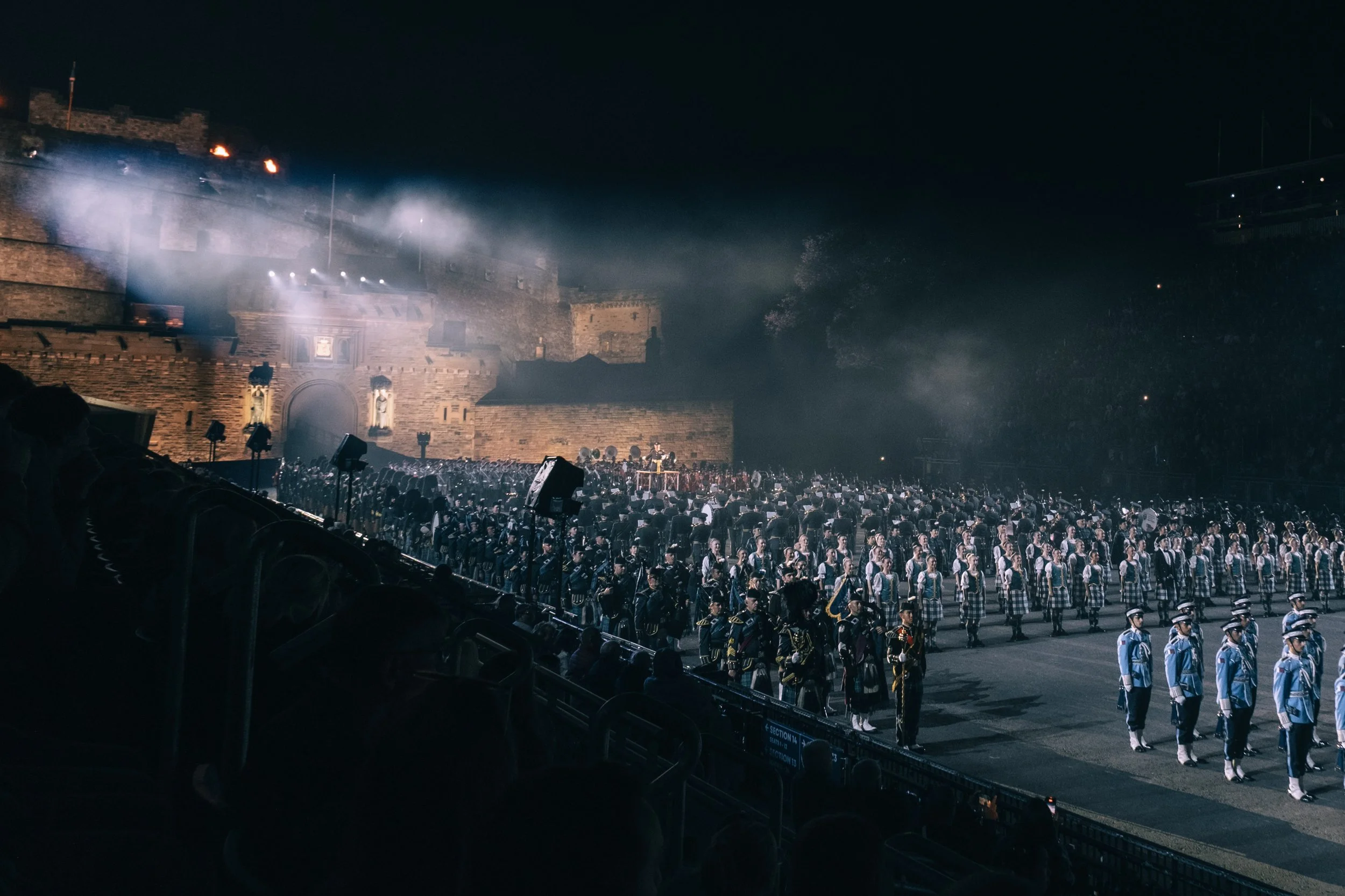 Nighttime military parade with soldiers in uniform standing in formation, illuminated by spotlights, with an ancient fortress wall in the background.