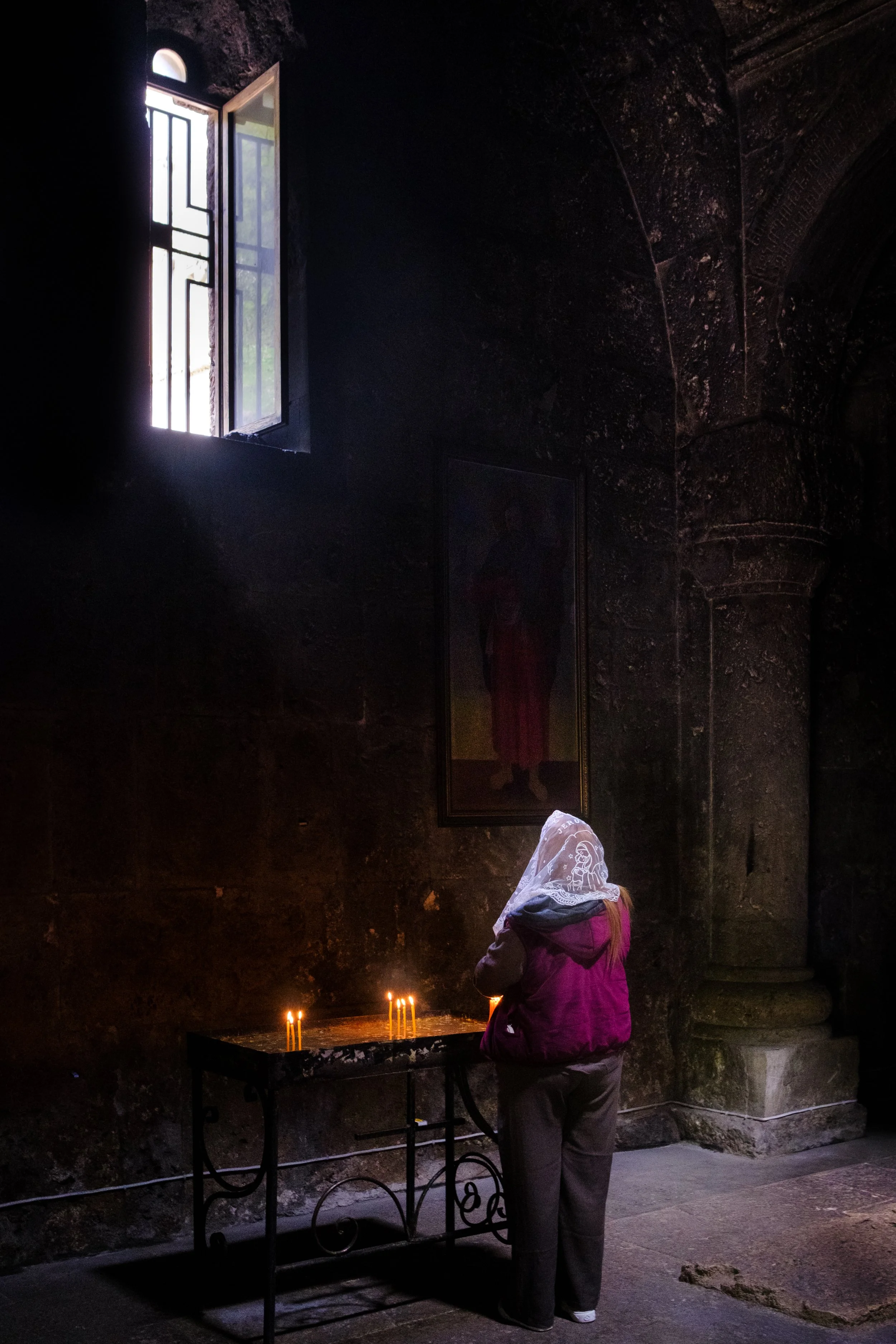 A woman with a white lace head covering, wearing a pink jacket and gray pants, kneeling before a row of lit candles on a black table inside a dark stone church or chapel, with a window letting in natural light, and framed art or icon on the wall.