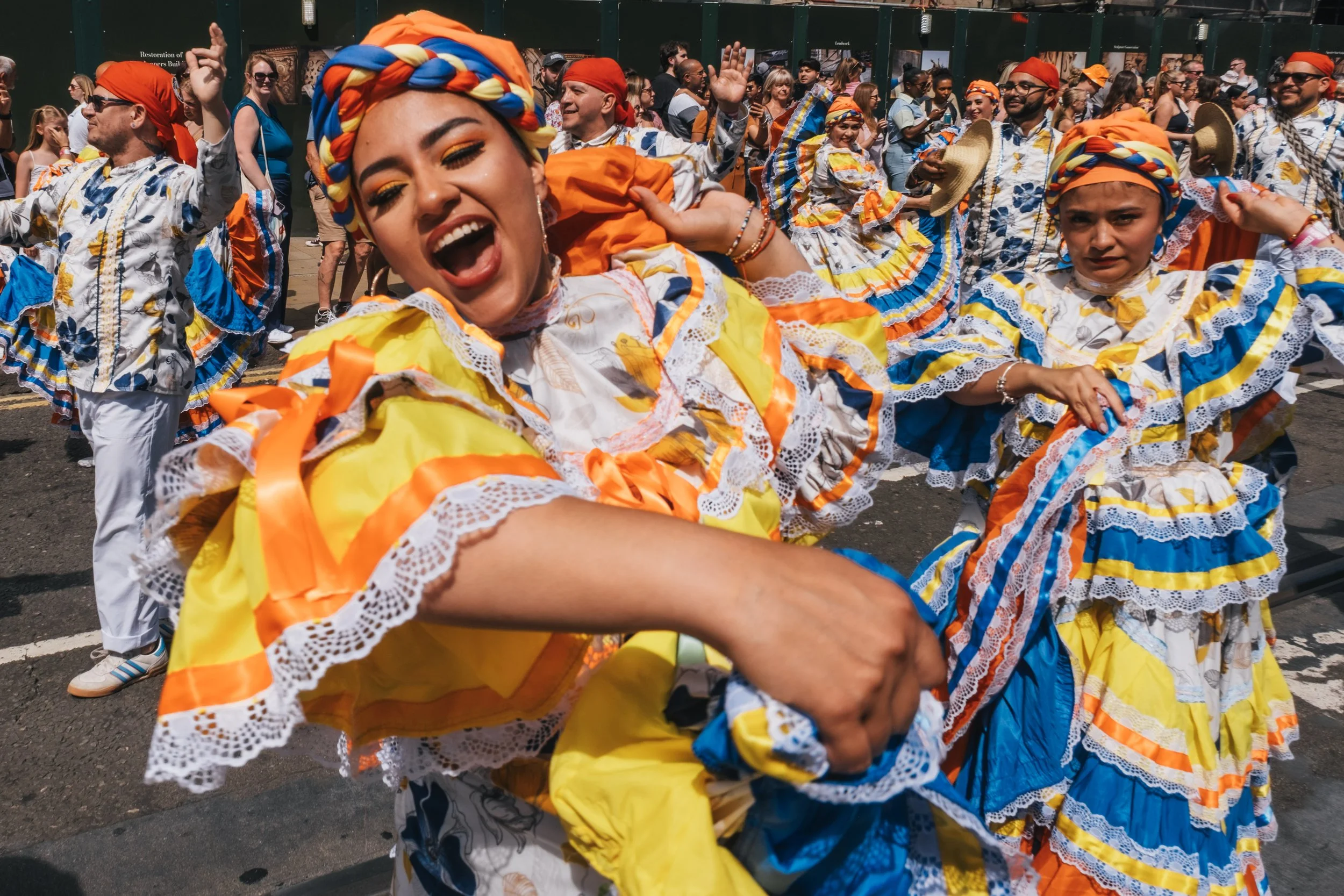 People wearing colorful traditional dresses and headscarves dancing and celebrating outdoors at a festival or parade.