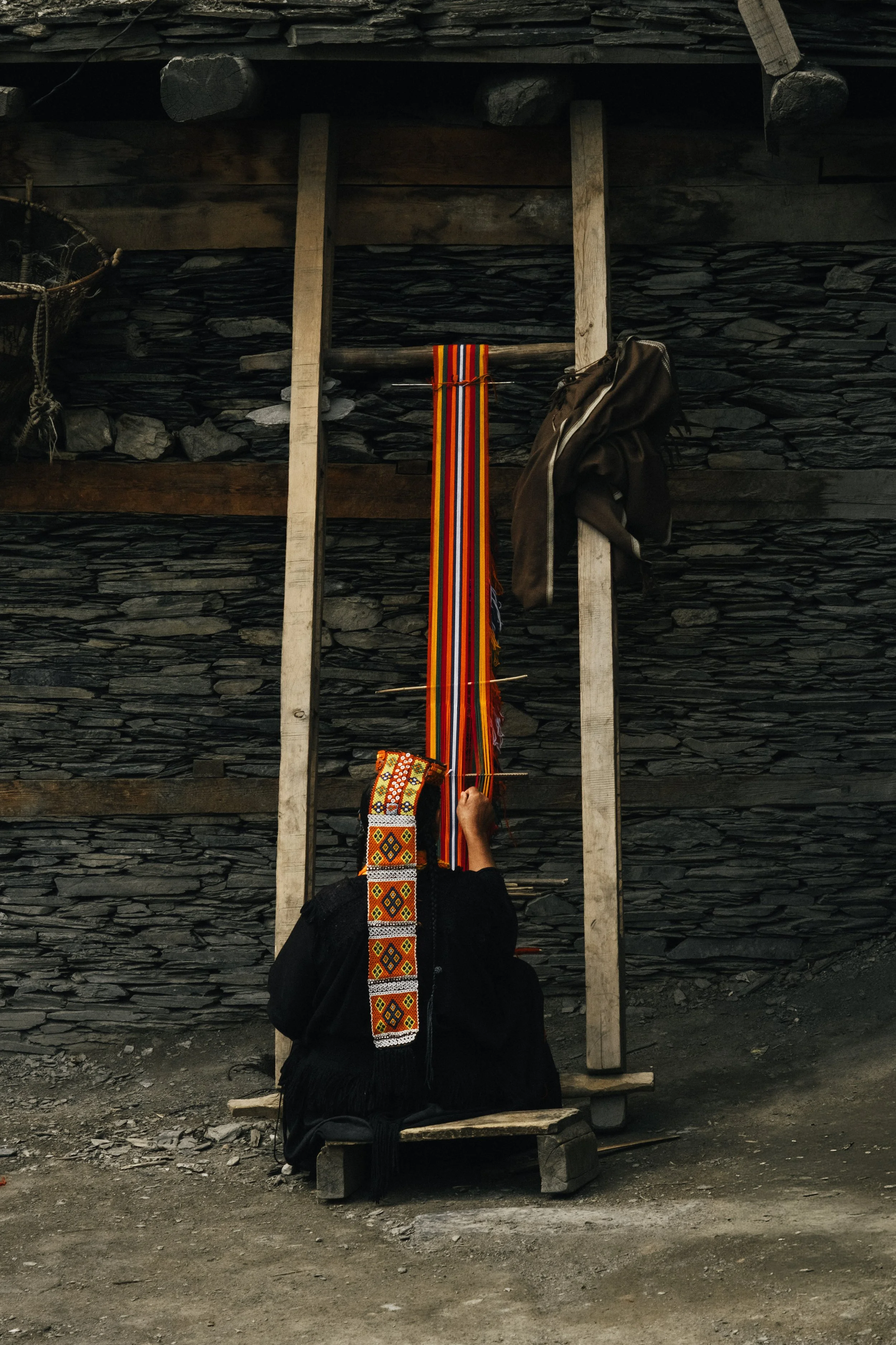 A person kneeling in front of a wooden frame, weaving colorful traditional fabric with a focus on intricate patterns, with a dark stone wall in the background.