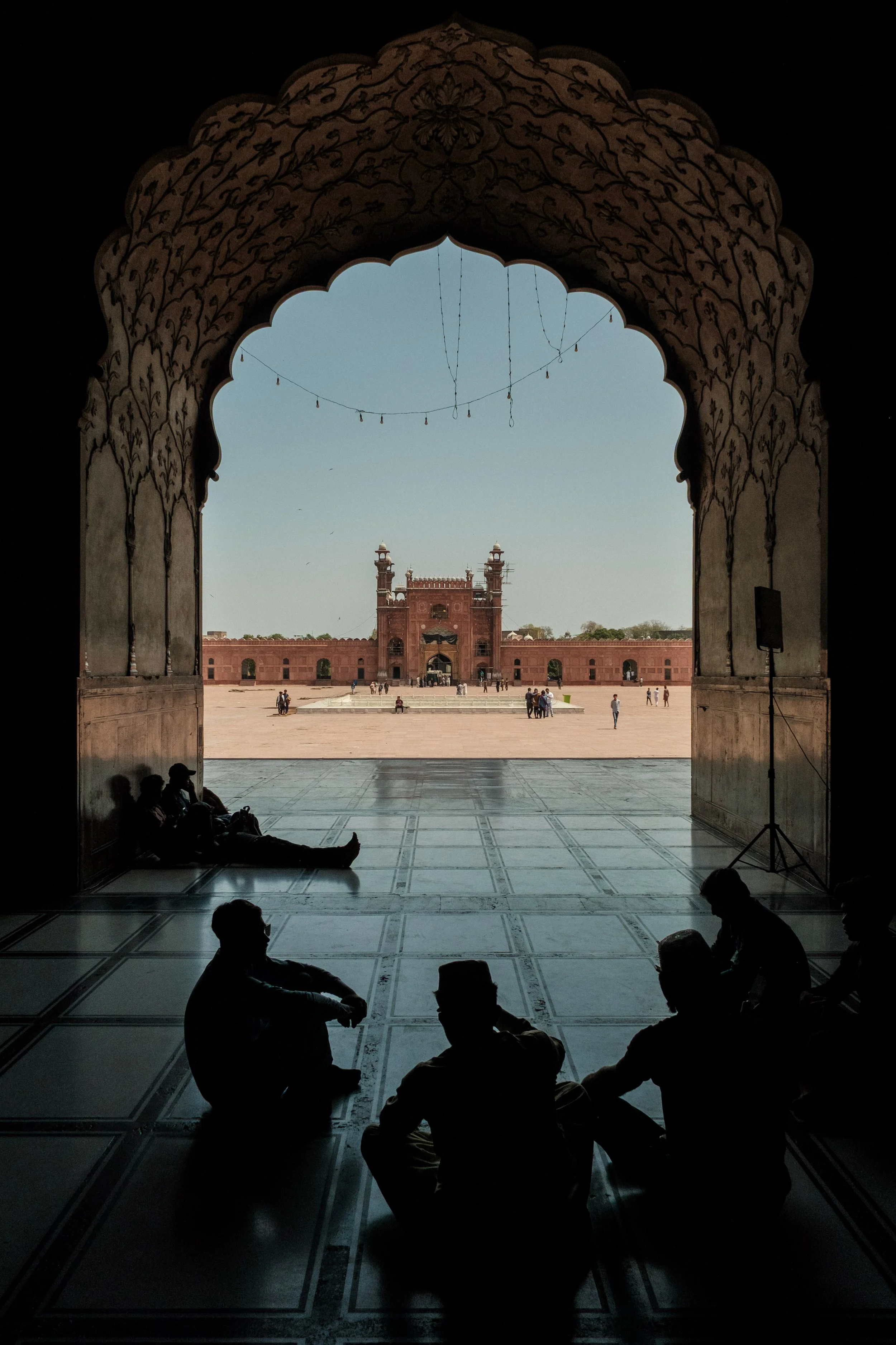 People sitting on the floor inside an archway, looking outward at a large red fort and open courtyard with a clear sky above.