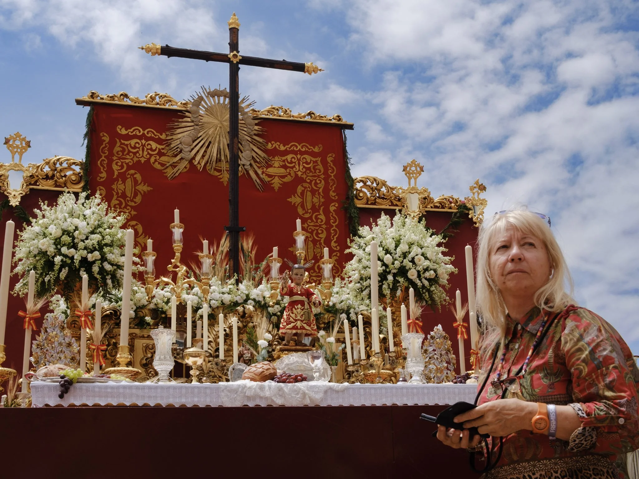 Religious procession with a decorated float featuring flowers, candles, and religious statues under a cloudy sky, with a woman in a floral dress standing nearby.