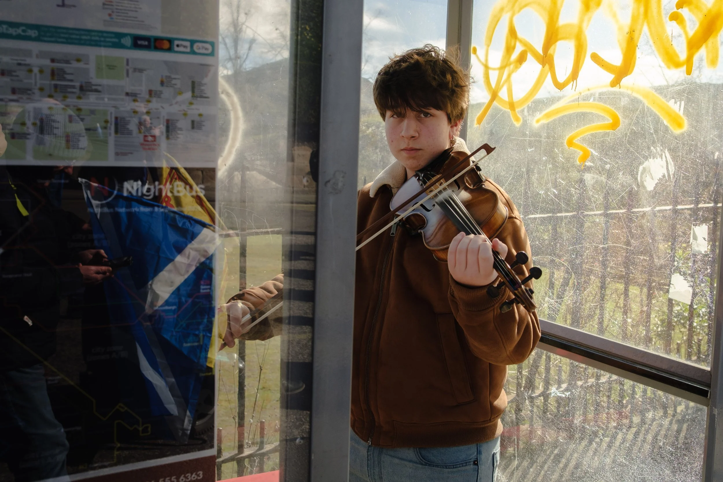 A young man with brown hair playing a violin inside a bus shelter, with reflections of people and trees visible through the glass.
