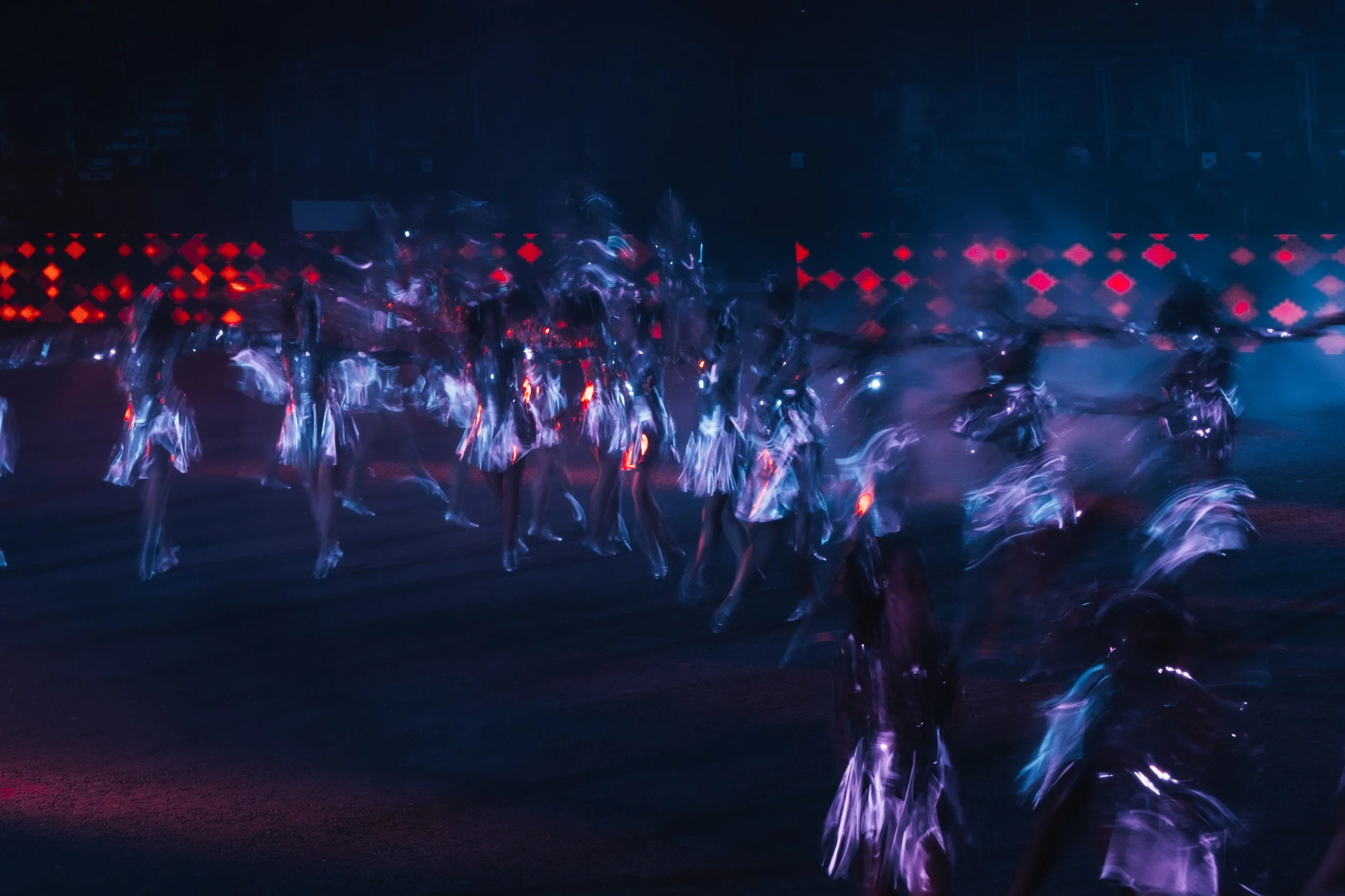 A group of people with illuminated outfits and reflective surfaces creating a glow, dancing or performing in front of a digital LED display with red geometric patterns at night.