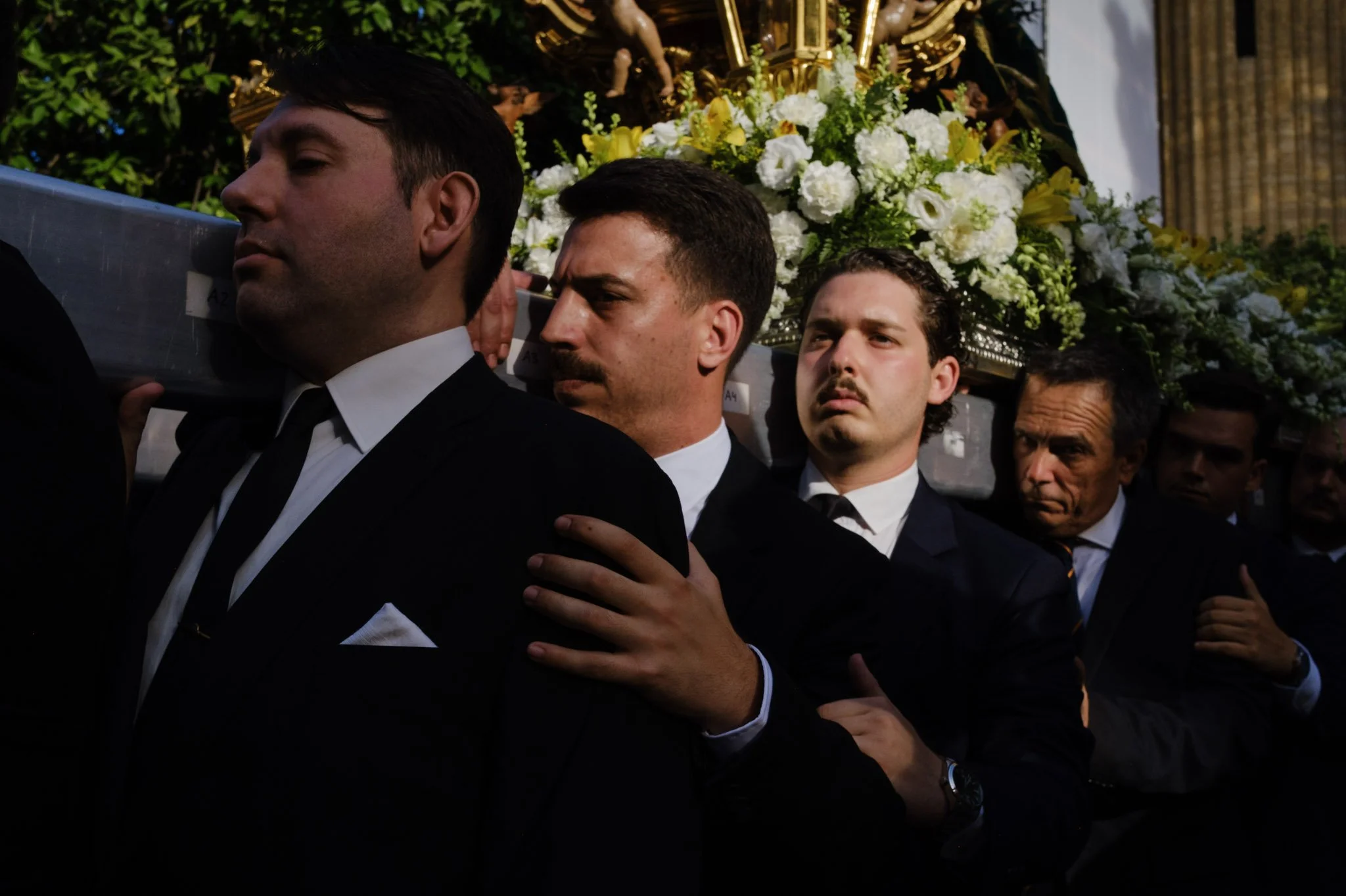 Men dressed in black suits carrying a religious statueadorned with white and yellow flowers.