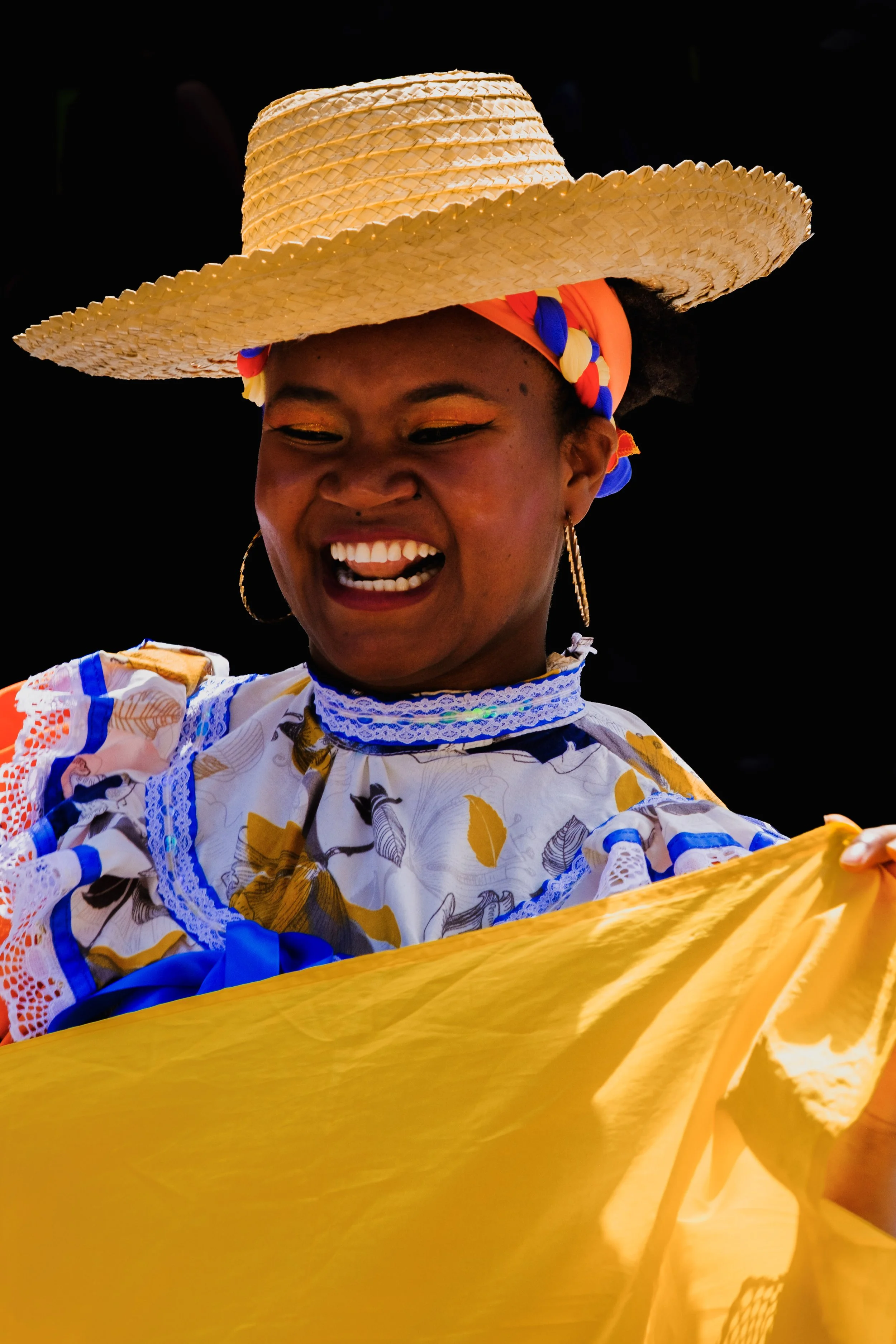 Woman wearing a wide-brimmed straw hat and colorful traditional dress, smiling and dancing during a cultural celebration against a black background.
