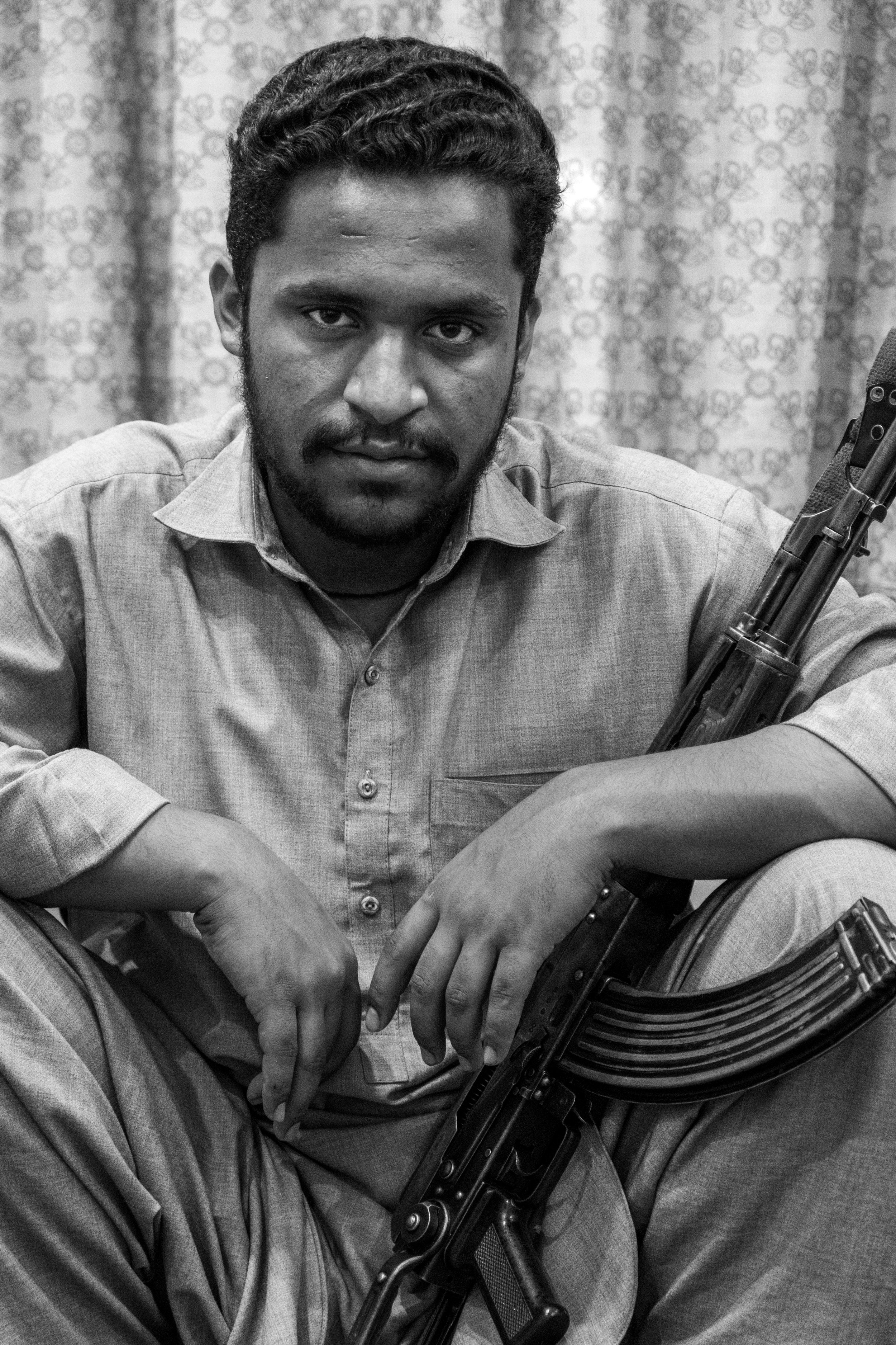 A man with dark hair and a beard sitting with his arms crossed on his knees, holding an automatic rifle, in front of a patterned curtain.