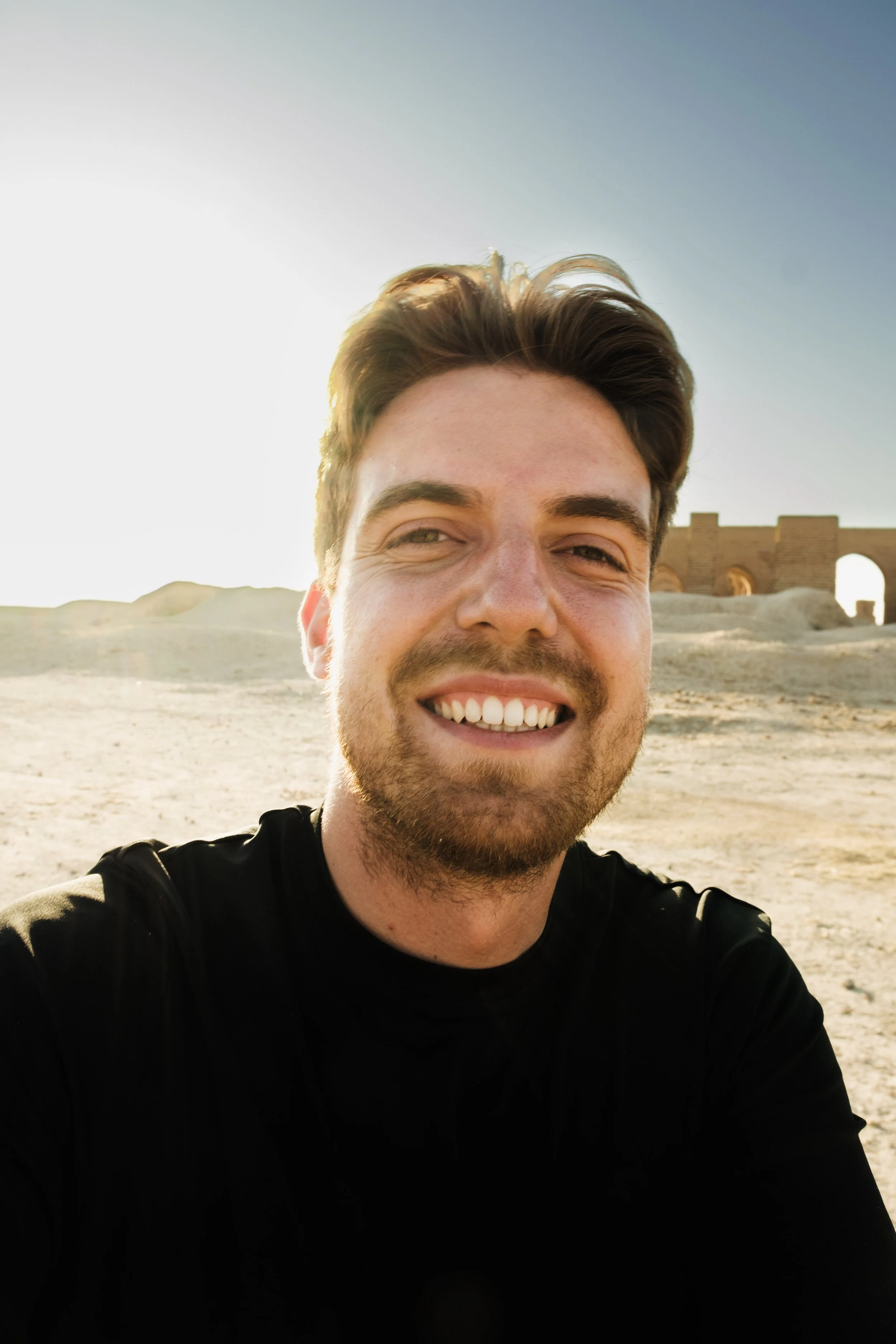 A smiling man with brown hair and a beard taking a selfie outdoors during sunset, with historical ruins in the background.