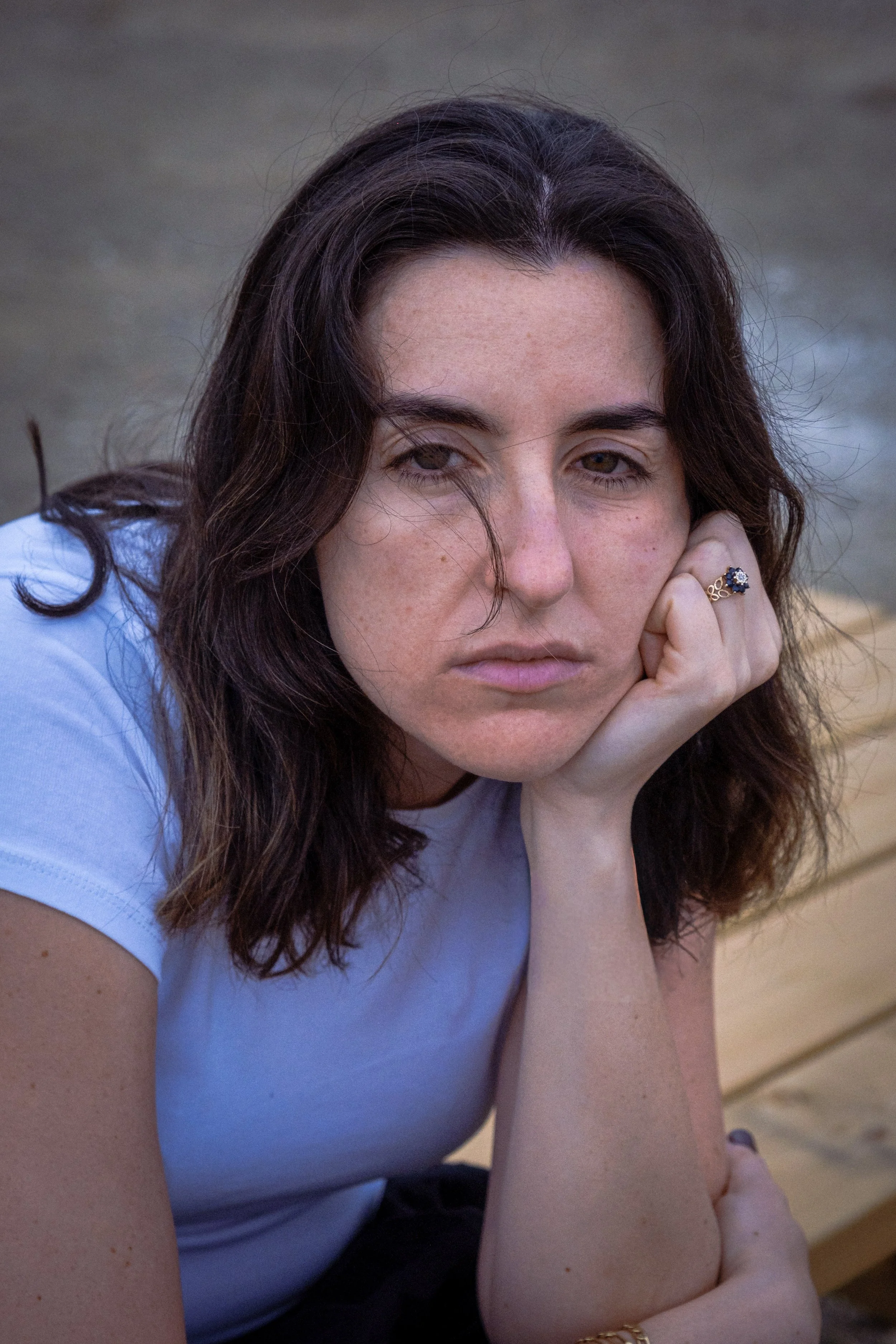 Close-up photo of a woman with brown hair, resting her head on her hand, wearing a white t-shirt, with a sandy beach background.