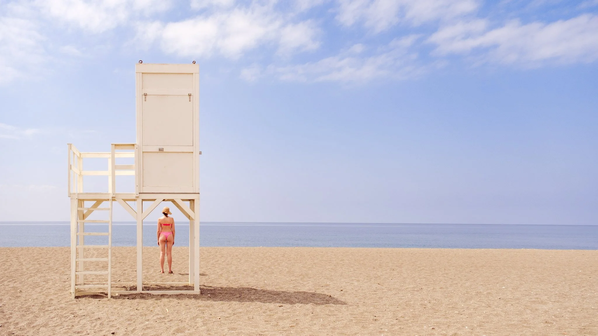 A person in a pink swimsuit standing on the sandy beach beneath a white lifeguard tower, with the ocean and sky in the background.