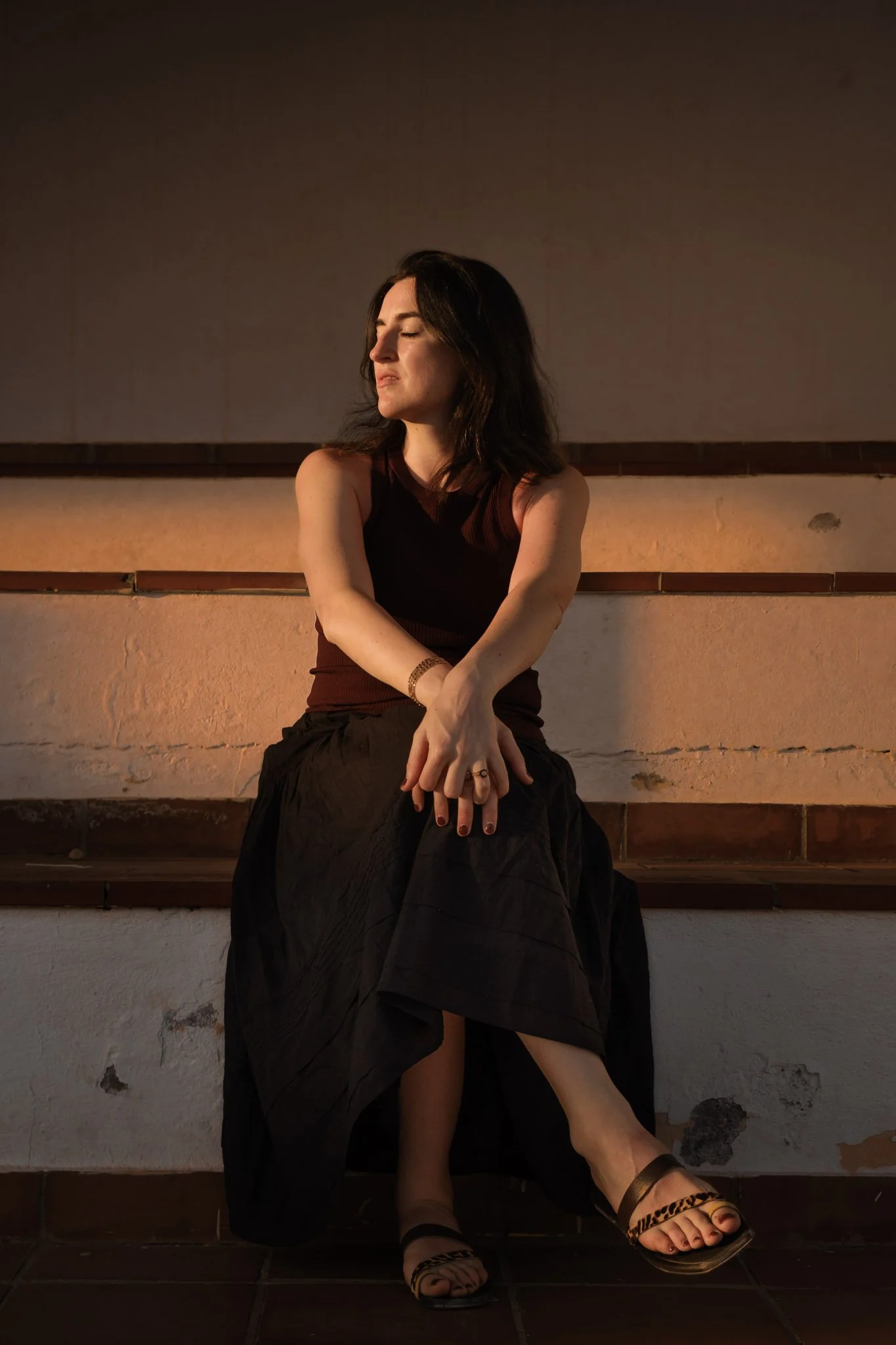 A woman with dark hair sitting on a bench against a wall, with her eyes closed, wearing a sleeveless black top and a long black skirt, posing with sunlit side lighting.