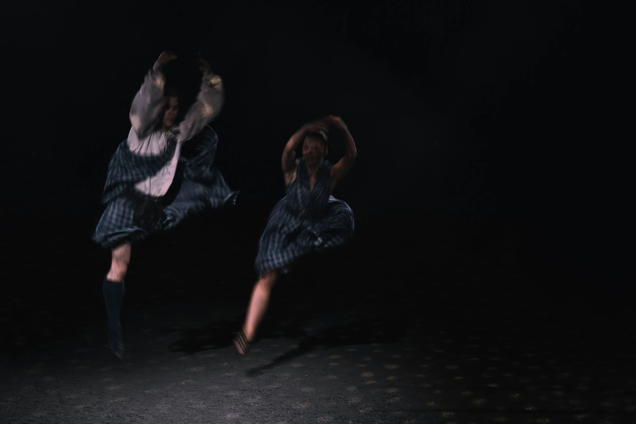 Two dancers performing a kick on a dark stage, wearing matching plaid costumes.
