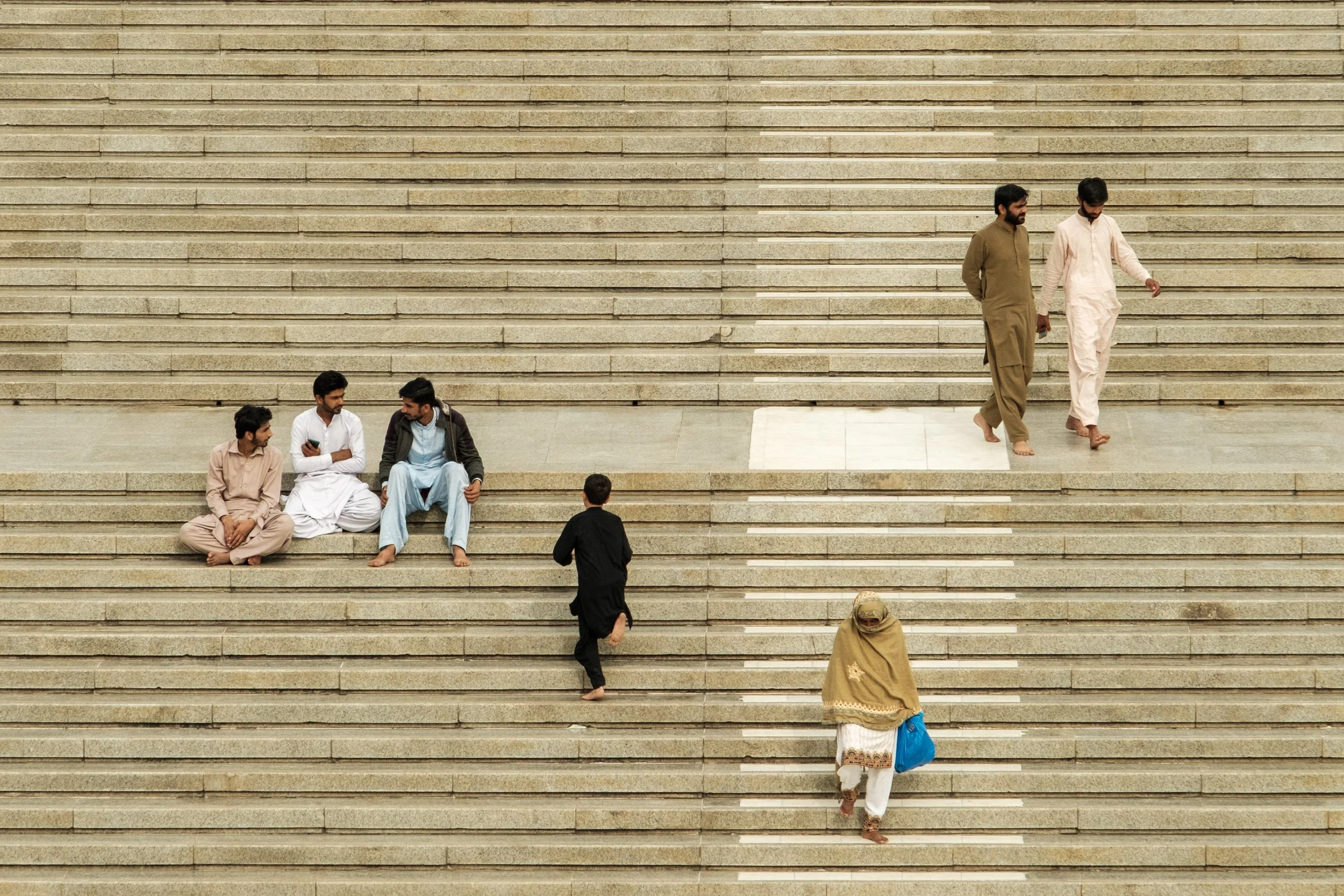Several people walking and sitting on a large set of stairs made of light-colored stone, with some sitting and some walking, wearing traditional and casual clothing.