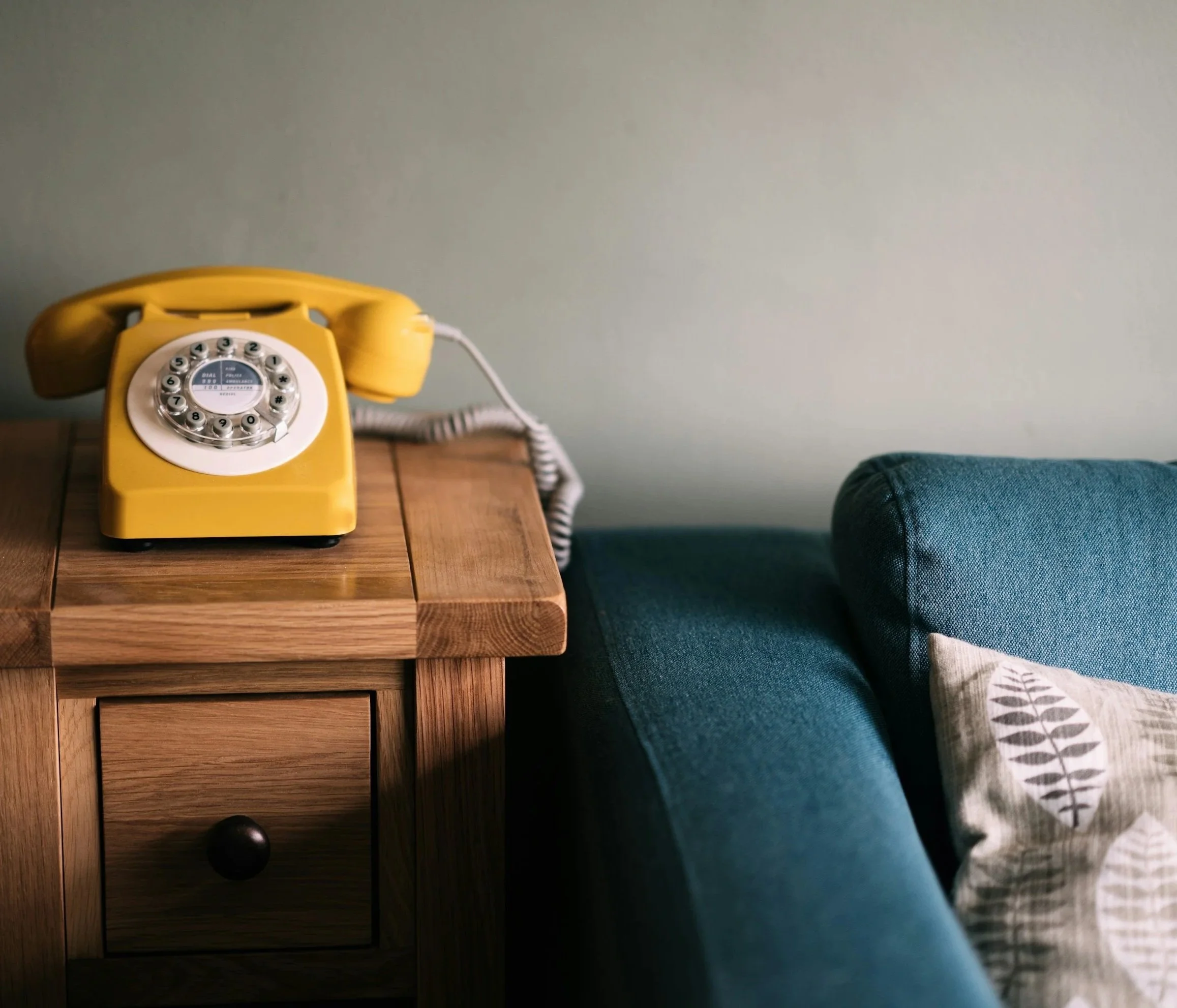 Yellow rotary telephone on a wooden nightstand next to a bed with a blue blanket and patterned pillow.