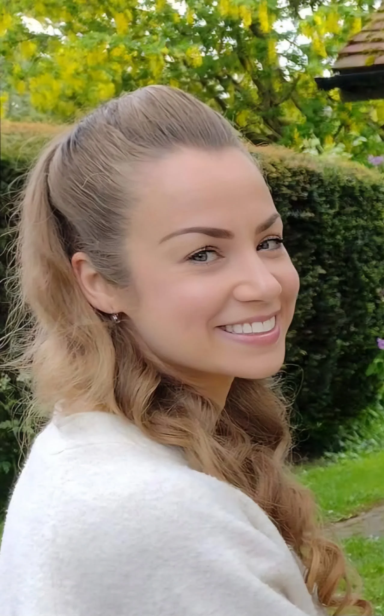 A woman with blonde hair, smiling outdoors with greenery in the background.