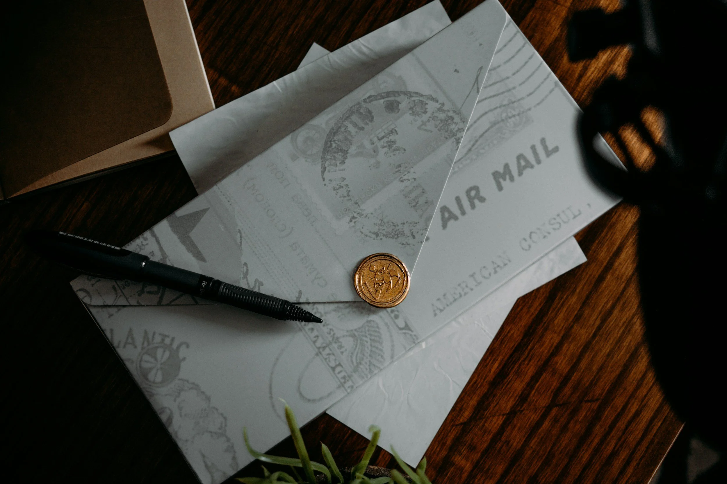 Close-up of a wooden tabletop with scattered envelopes, a black pen, a gold coin, and a partially visible black object, with some green plant stems in the foreground.