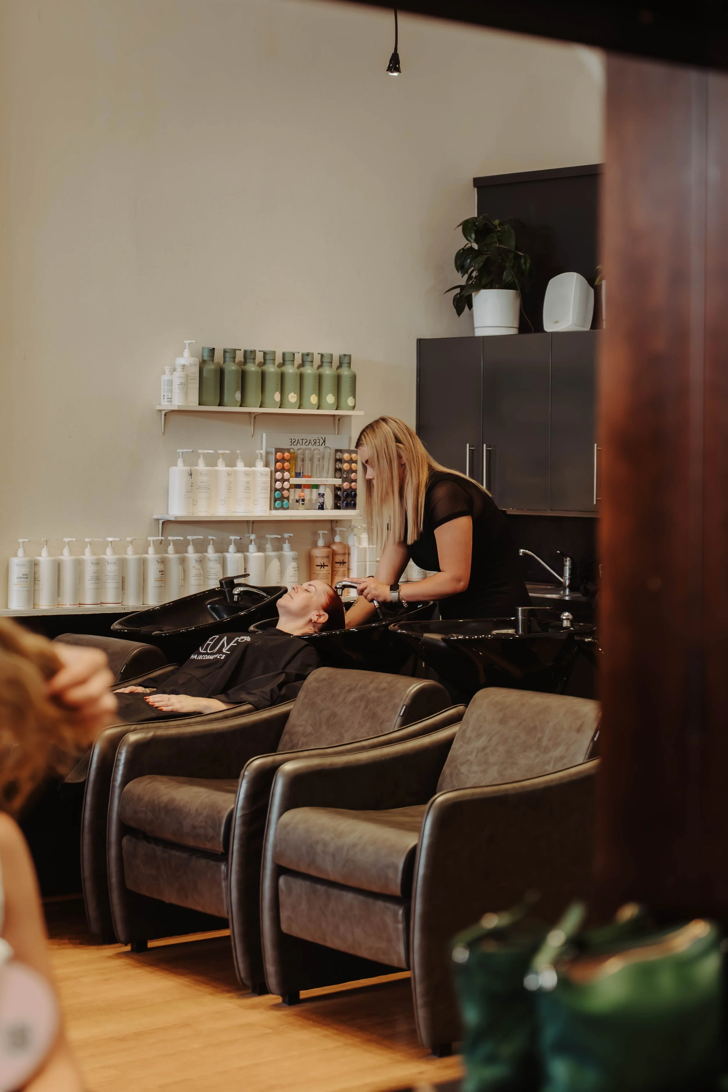 A person is getting their hair washed at a salon while another person is reclining in a salon chair with their eyes closed. The salon has black shelves with hair care products and a potted plant on top.