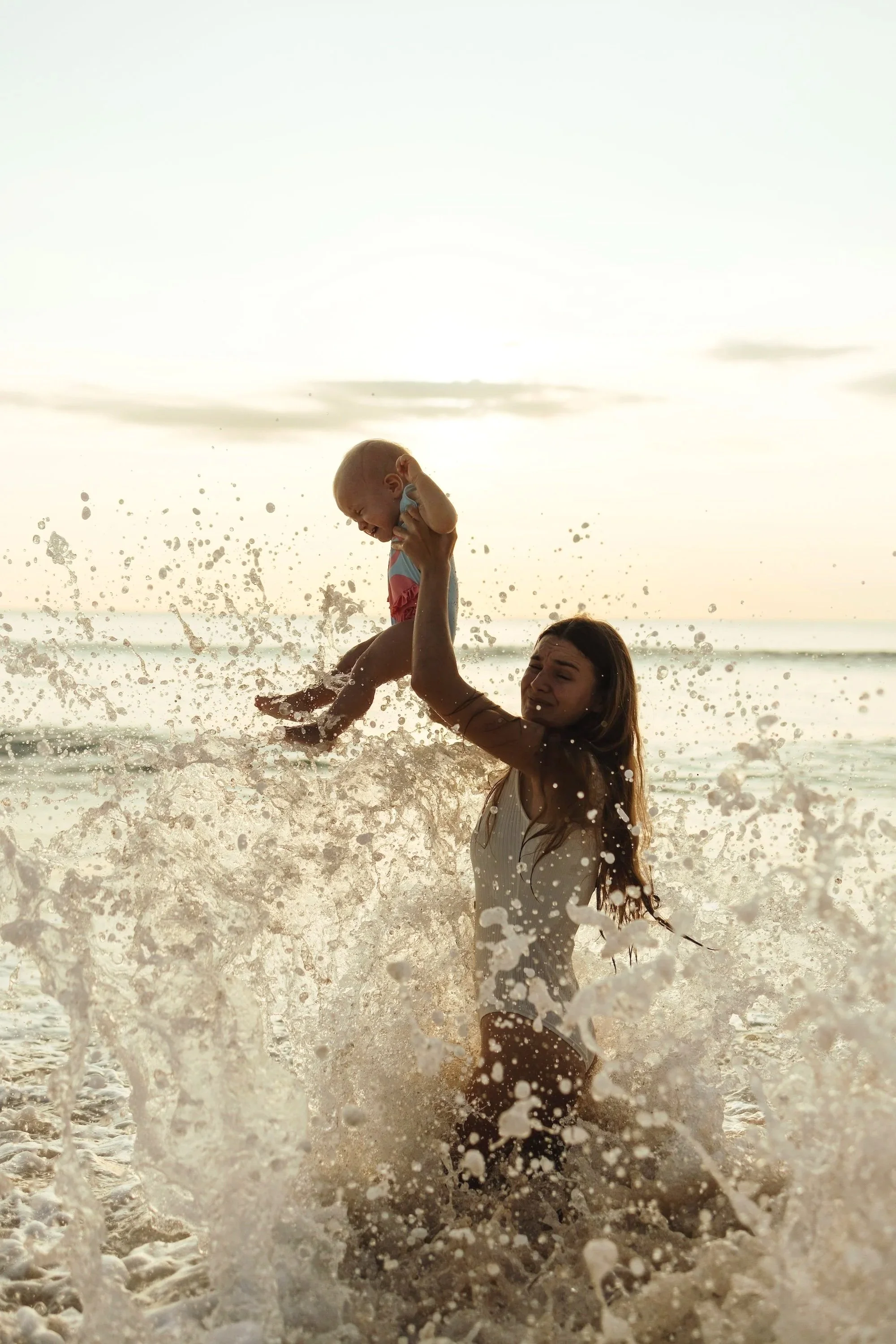 young woman lifting baby in the waves at sunset