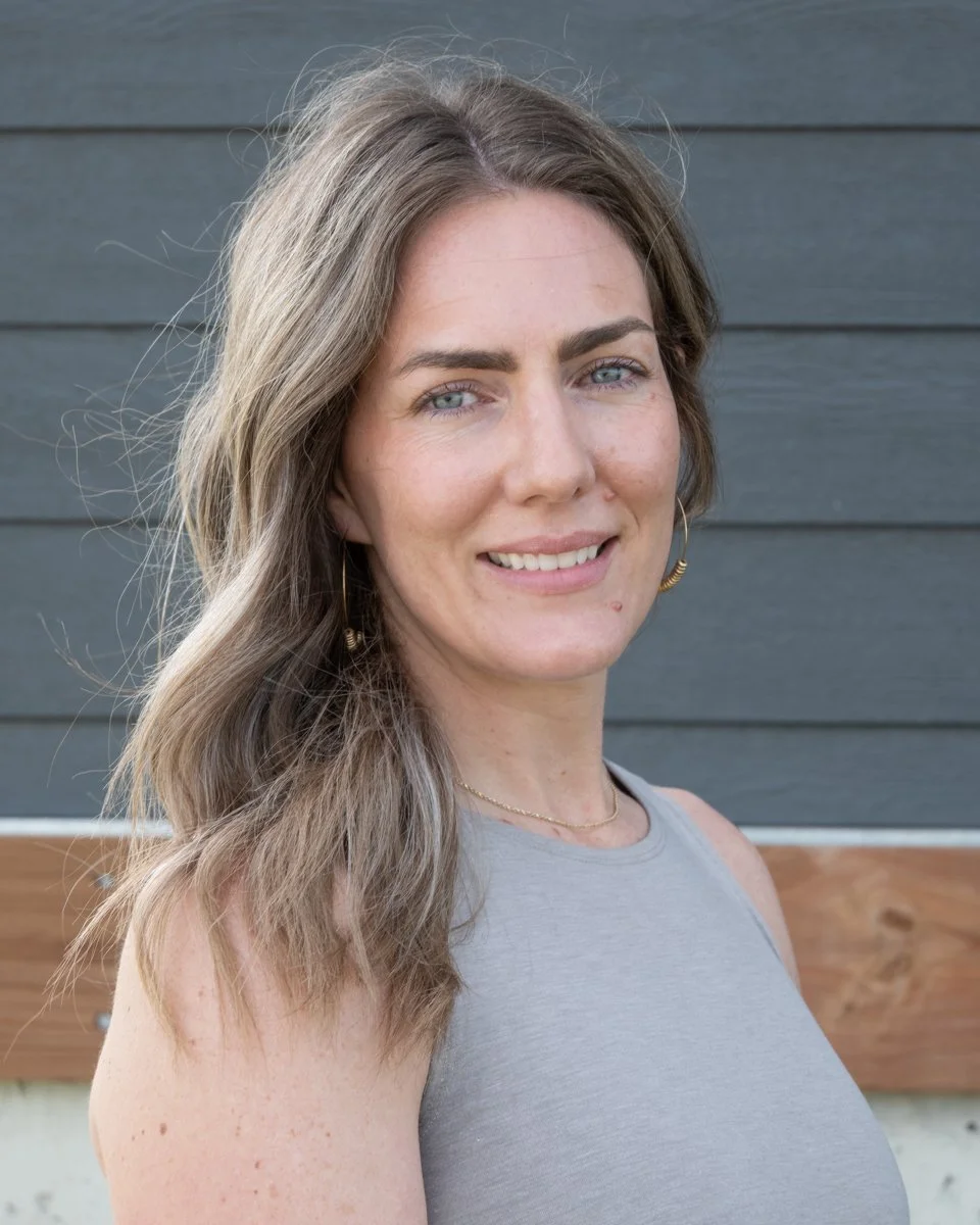 A woman with long, wavy brown hair and blue eyes, smiling, wearing a sleeveless gray top, earrings, and a necklace, standing outdoors against a gray wooden wall.