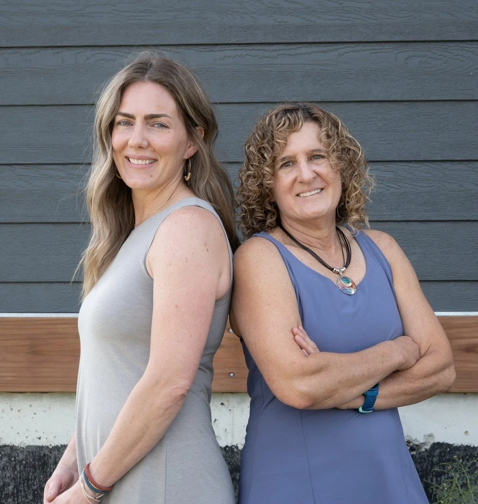 Two women standing back-to-back outdoors, smiling, with a gray and wood-paneled wall in the background.