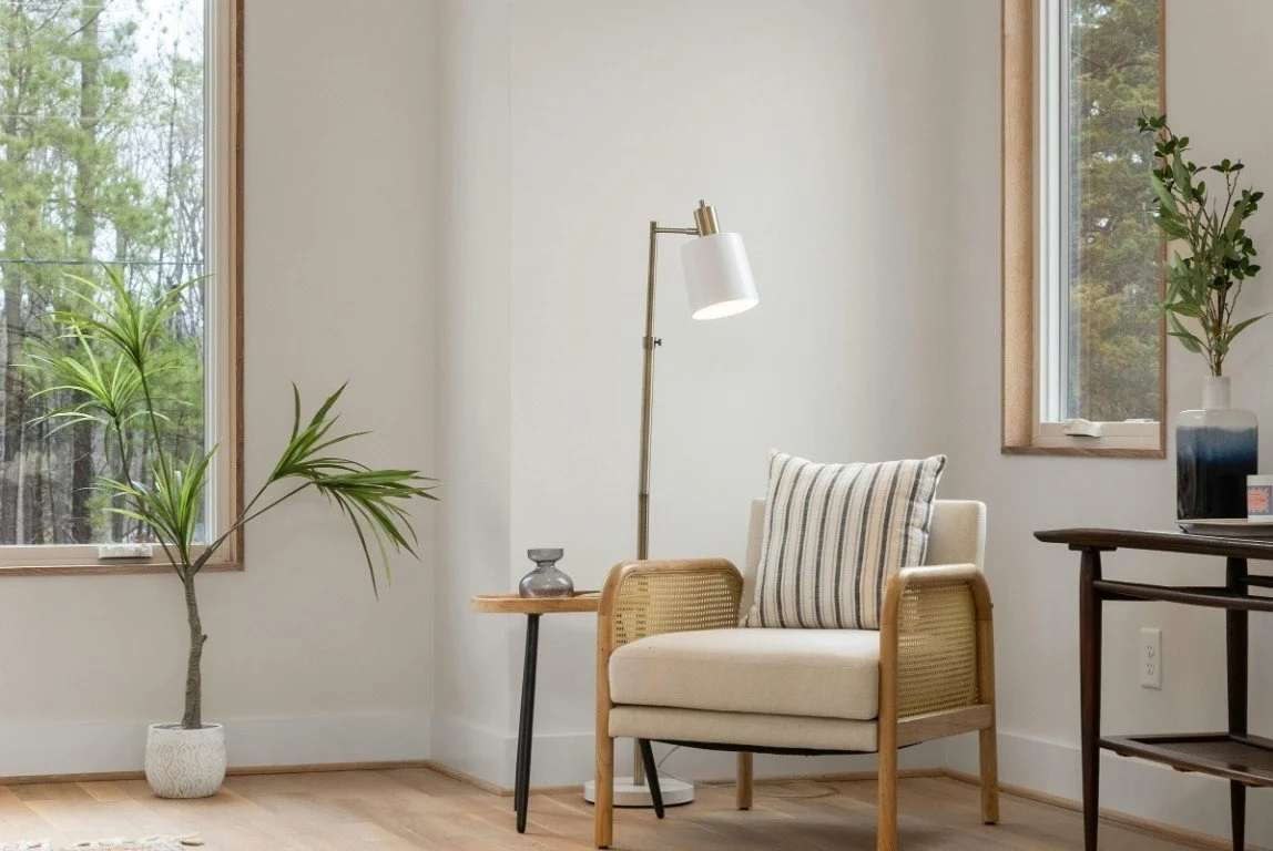 A cozy corner of a room with a beige armchair, a striped pillow, a wooden side table with a small decorative vase, a tall floor lamp, and a window with greenery outside.