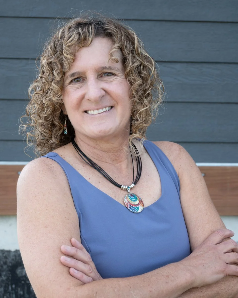 A woman with curly blonde hair smiling and crossing her arms, wearing a blue sleeveless top and a colorful circular pendant necklace, standing in front of a gray wooden wall.
