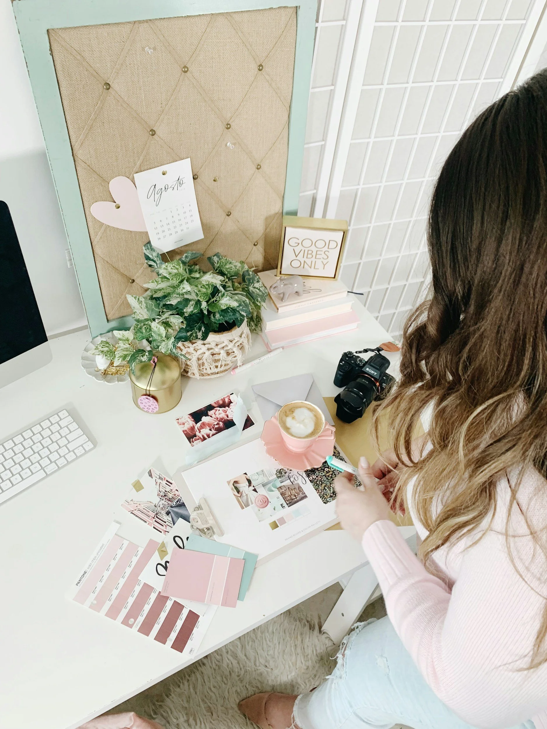 A woman sitting at a white desk with various color swatches, magazines, and design materials, working amidst decor and motivational items such as a 'Good Vibes Only' sign, a potted plant, and a calendar.