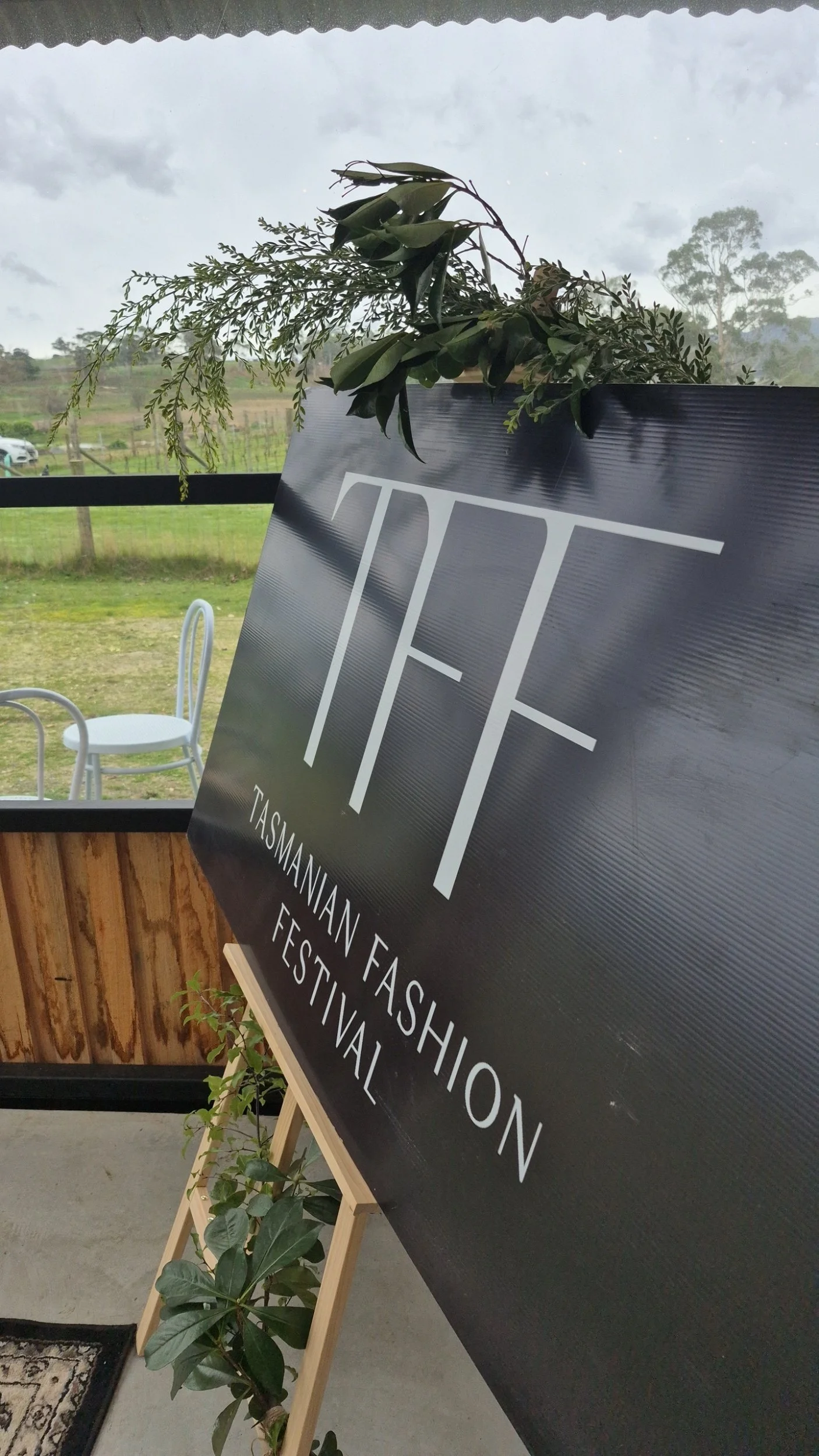 A black sign with white text advertising the Tasmanian Fashion Festival, placed on an easel in a room with wooden paneling. A plant is next to the sign. In the background, there are outdoor views through a window, including trees, a cloudy sky, and a white chair.