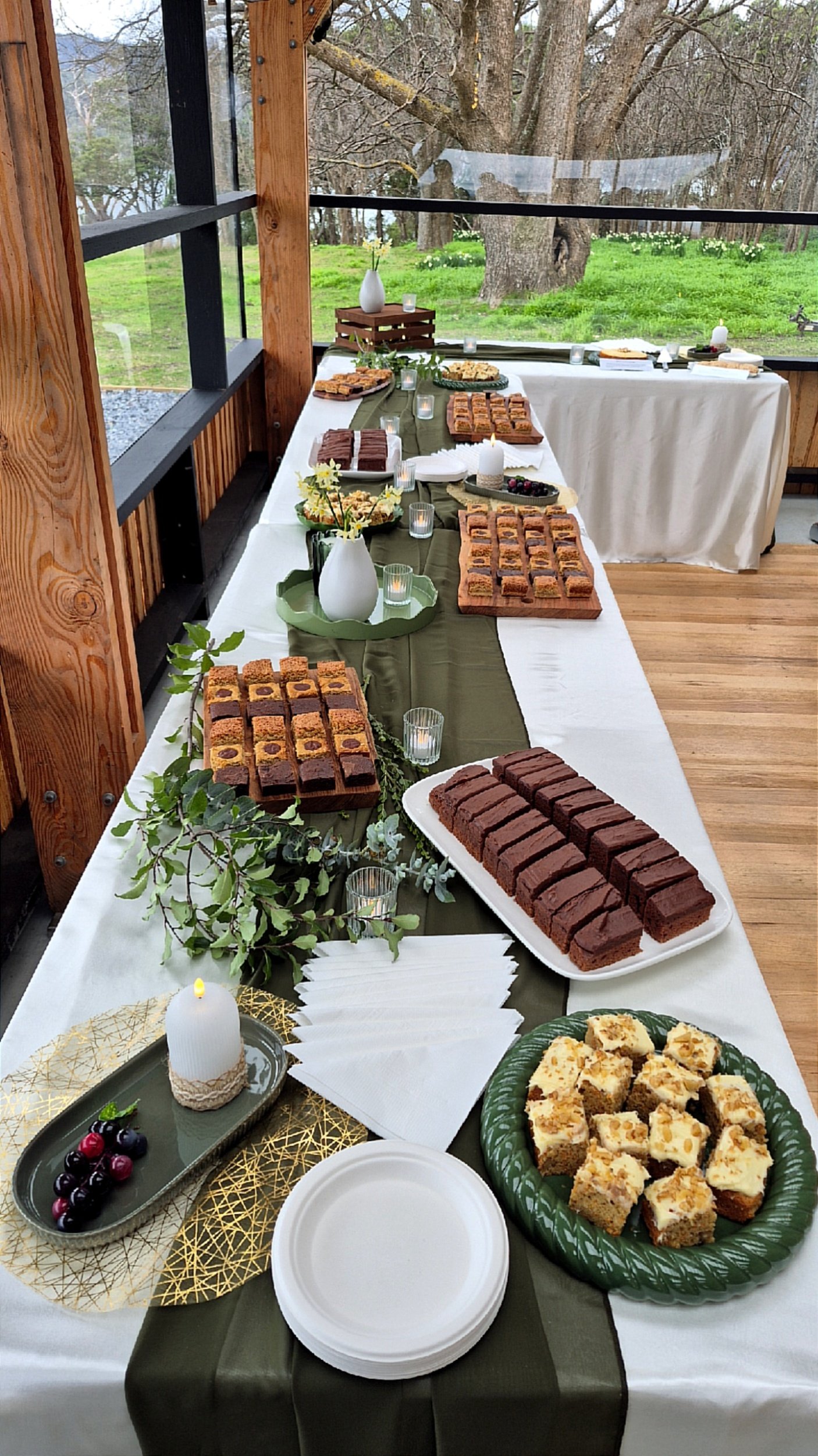 A long table set with various dessert bars, cookies, cupcakes, and fruit, decorated with green and gold accents, candles, and potted plants, outdoors with grass and trees in the background.