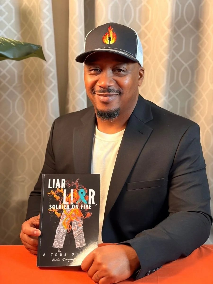 A man in a black blazer and white t-shirt wearing a black and gray cap with a flame and torch logo, sitting at a table in front of beige patterned curtains, holding a book titled 'LIAR LIAR, SOLDIER ON FIRE'.
