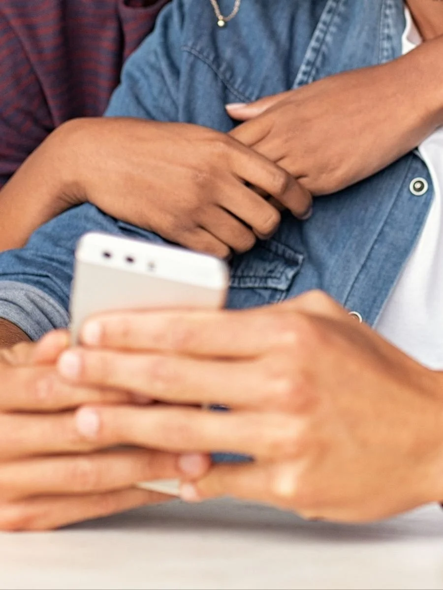 Two people, one with darker skin and one with lighter skin, sharing a moment with one person embracing the other while looking at a smartphone.