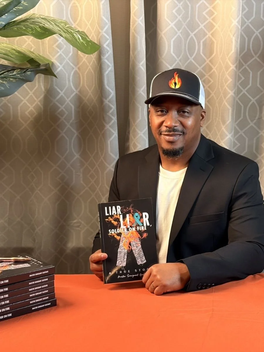 A man in a black blazer and white shirt is sitting at a table with a stack of books titled 'LIAR' and 'SOLDIER ON FIRE' in front of him. He is holding one of the books and smiling at the camera. He wears a baseball cap featuring a fire emoji with a flame in the shape of a person.